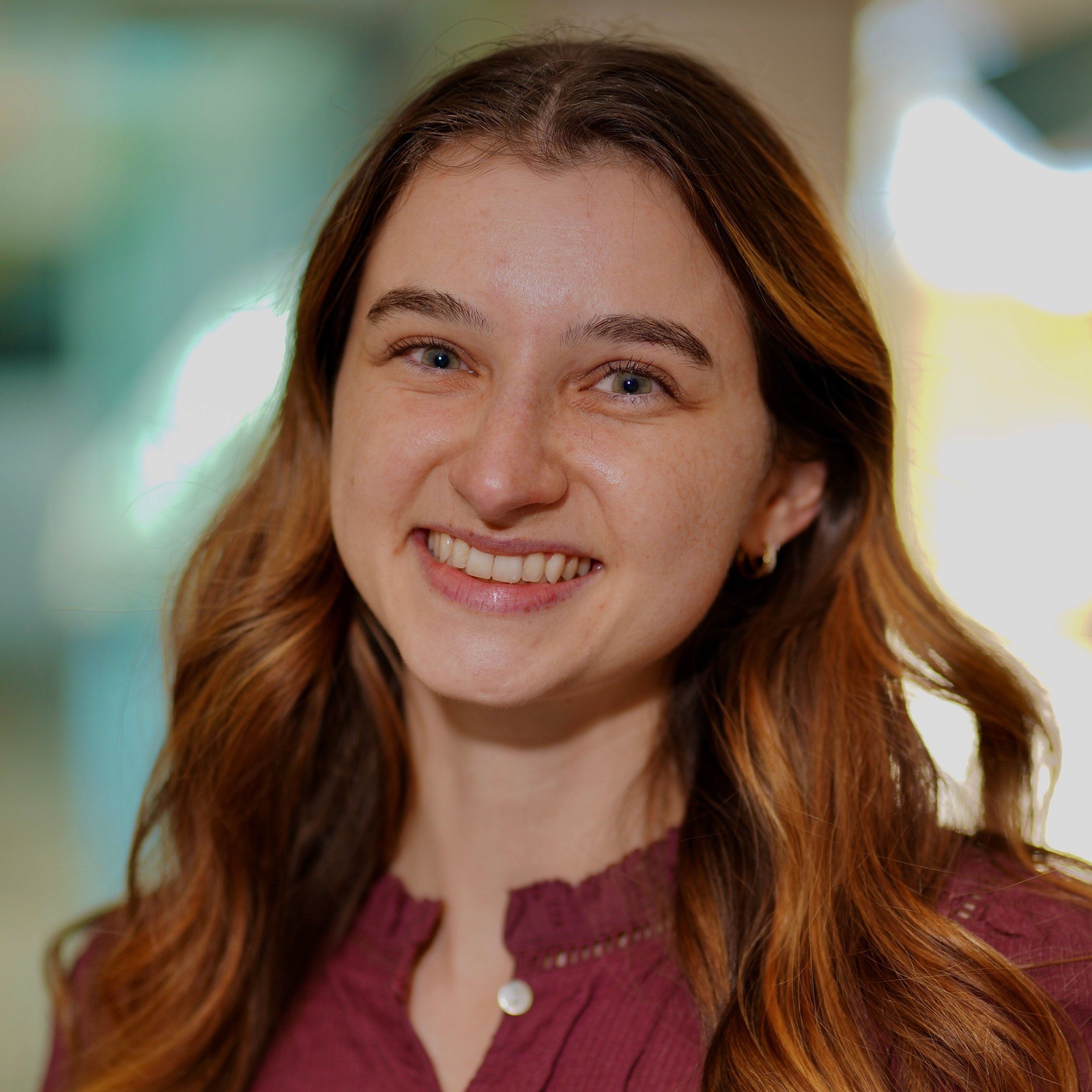 A young woman with long, wavy brown hair smiling at the camera against a plain light-colored background.
