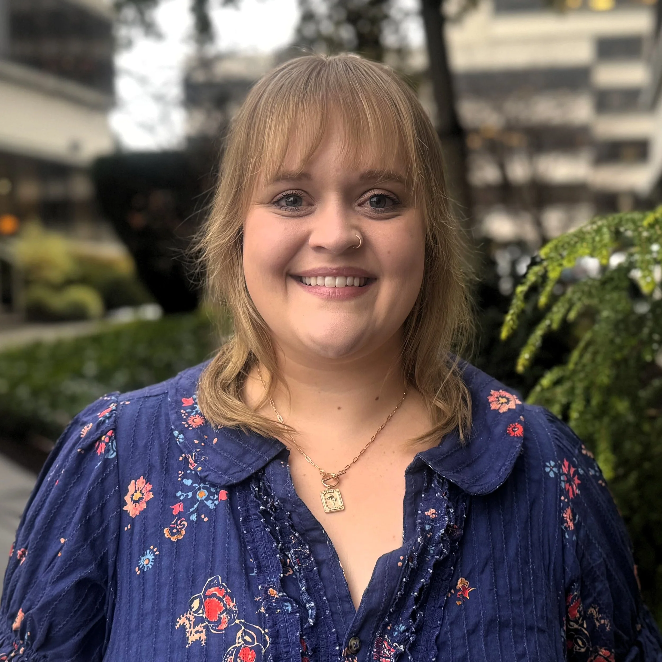 A woman holding a small brown and white dog outdoors, with green trees and cloudy sky in the background.