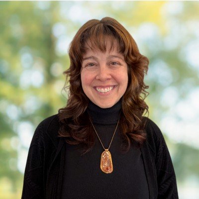 A woman with medium length brown hair and a bright smile, wearing a green top, poses against a dark grey background.