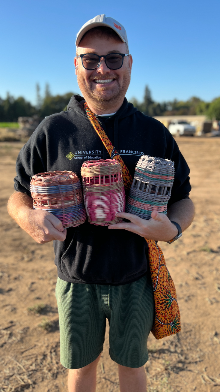 Smiling man wearing sunglasses, a baseball cap, and a University of San Francisco hoodie, holding three colorful woven baskets outdoors on a sunny day.