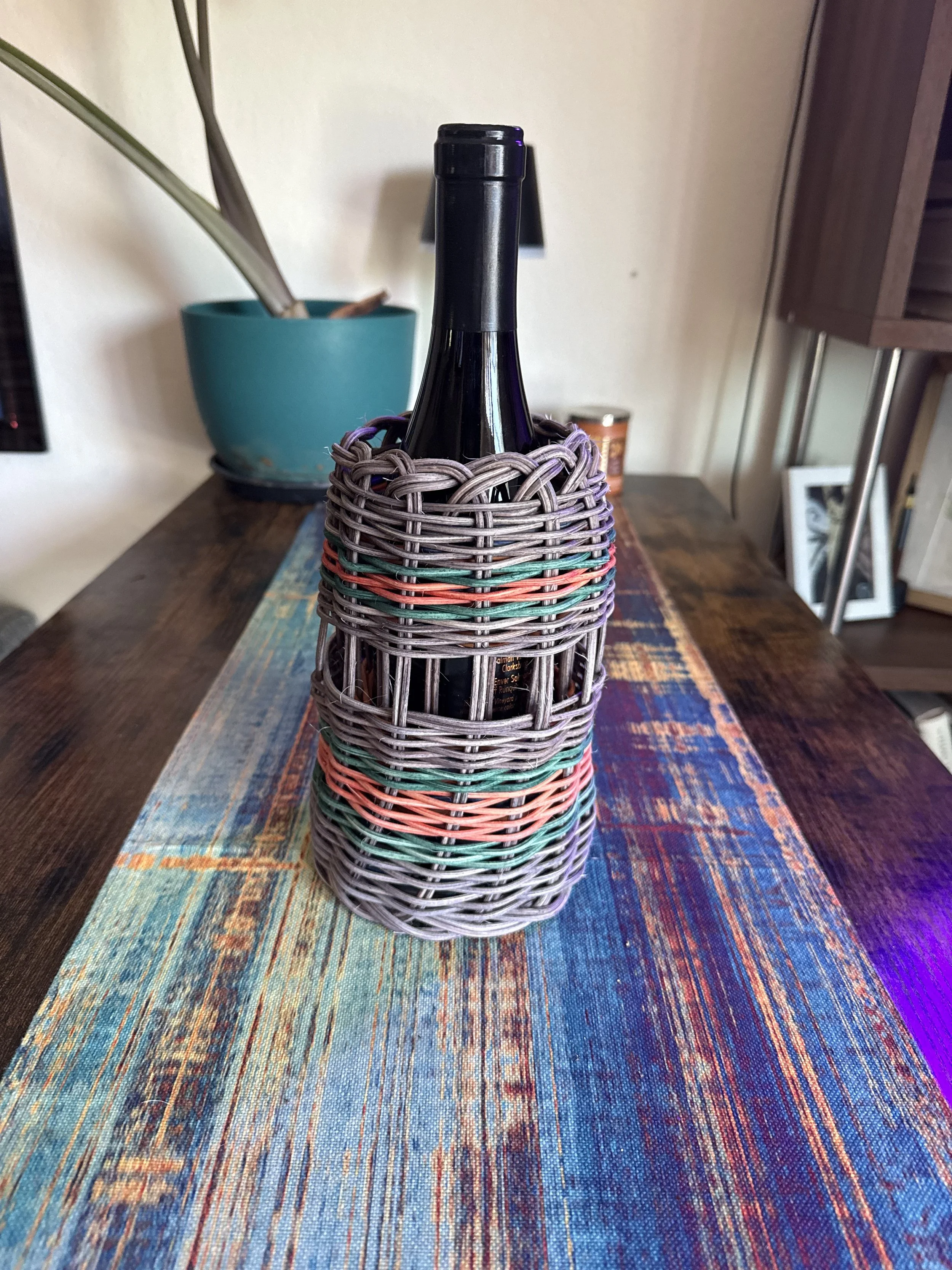 A woven bottle holder with a dark glass bottle inside, placed on a colorful fabric runner on a wooden table in a room with a large teal pot, a picture frame, and some shelves.