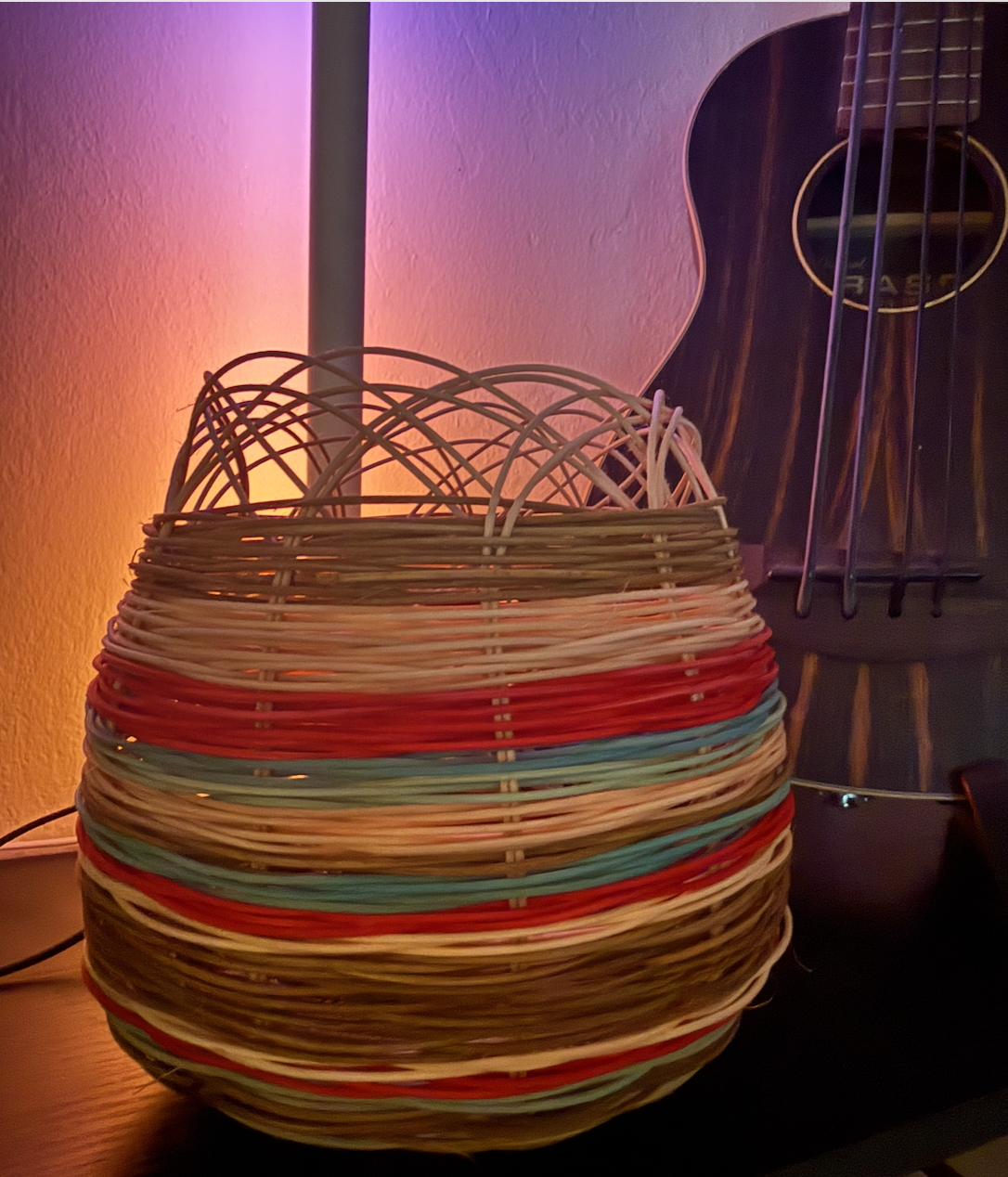 Colorful woven basket with a lamp or light source inside, multicolored with shades of brown, red, blue, and beige, and a guitar partially visible in the background.