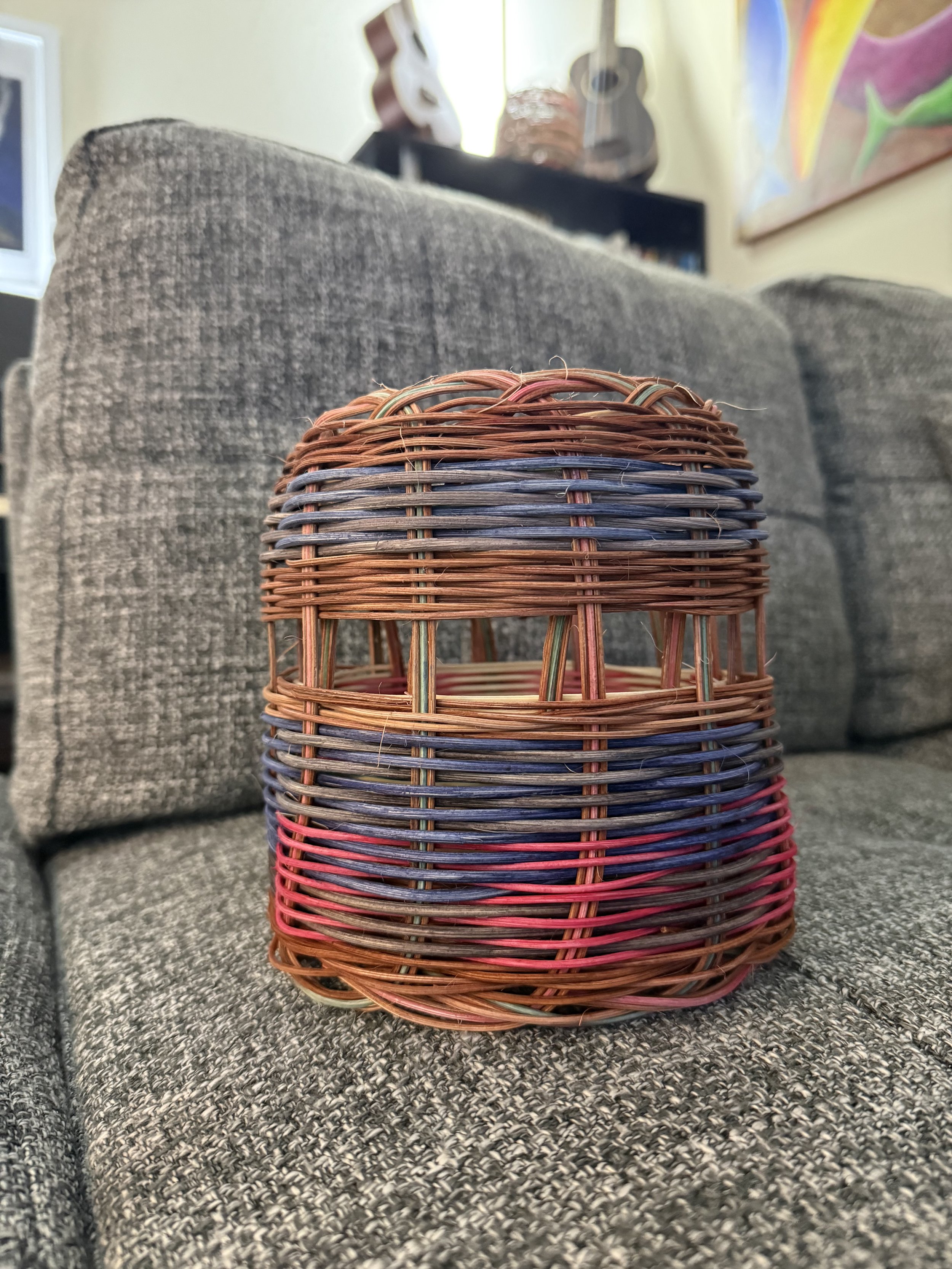 Close-up of a woven, multi-colored wicker basket on a gray sofa in a living room with wall decorations and a painting.