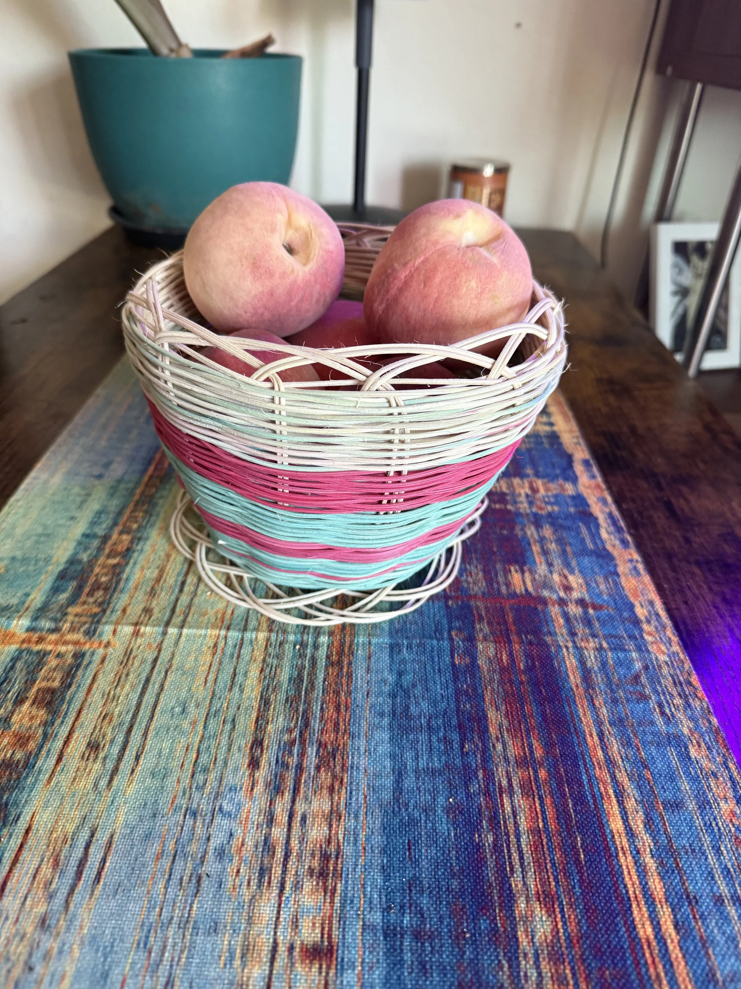 A woven basket with pink peaches sits on a colorful woven tablecloth, with a teal planter, a black lamp, a brown container, and a picture frame in the background.