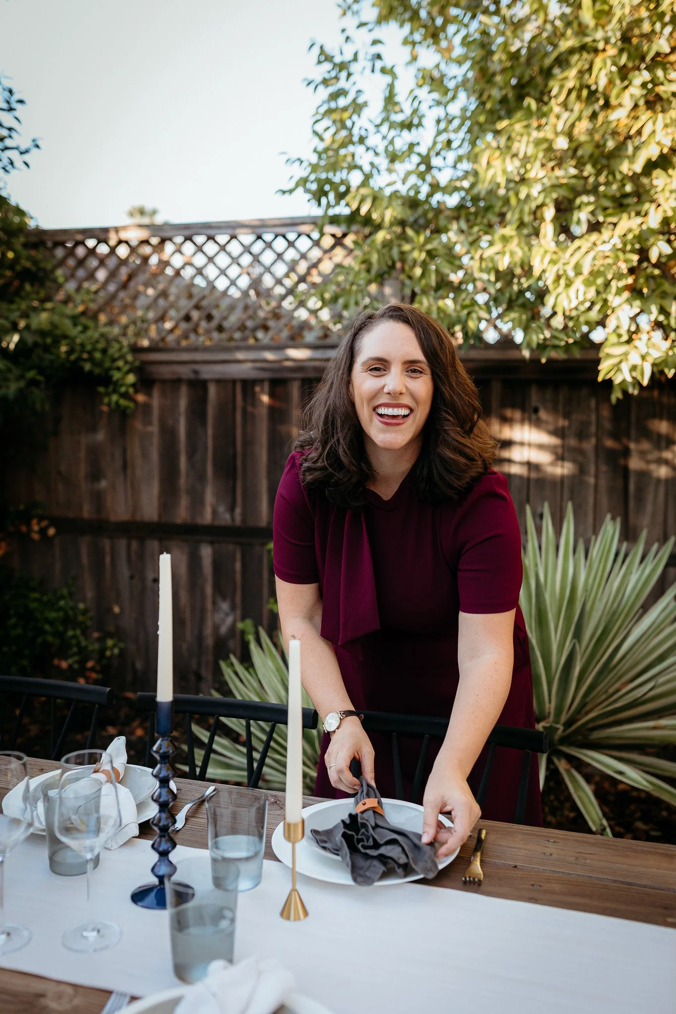 A woman with dark wavy hair, wearing a burgundy dress, is smiling while standing outdoors at a table set for a meal. There are candlesticks, glasses, and napkins on the table, with a wooden fence, green plants, and trees in the background.