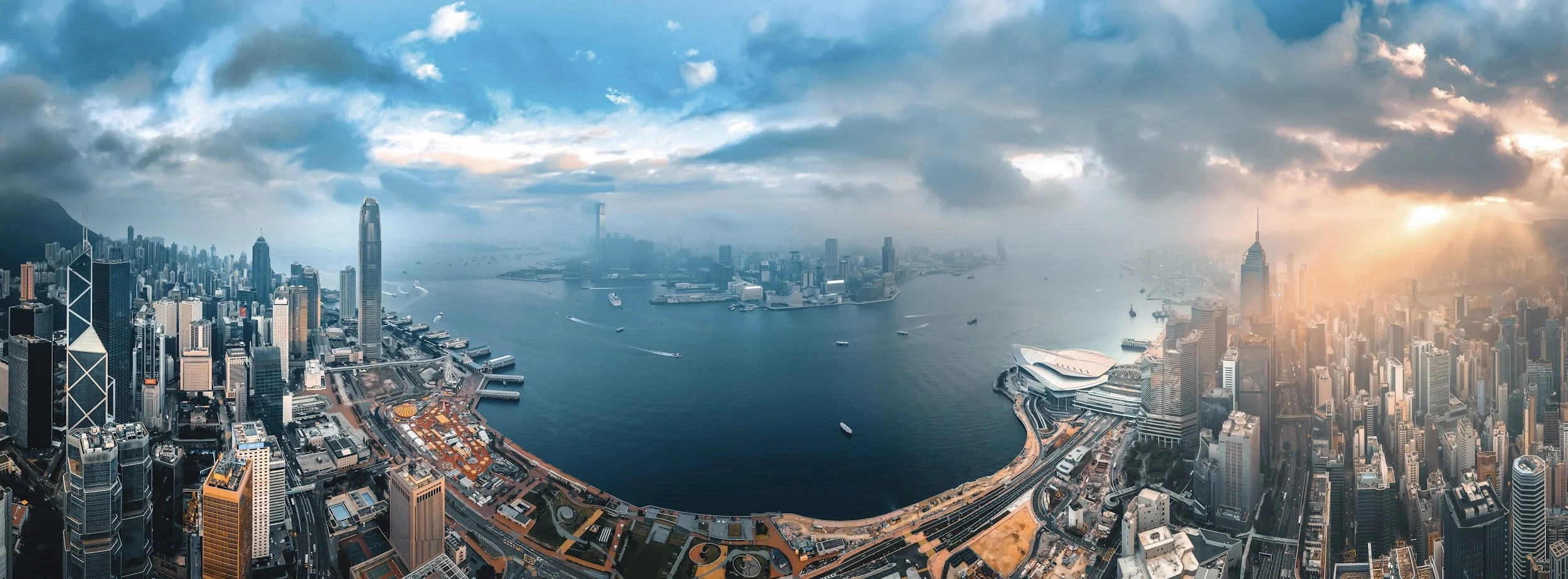 Aerial view of a city skyline with skyscrapers, harbor, and ships, showing a sunset with dramatic clouds.