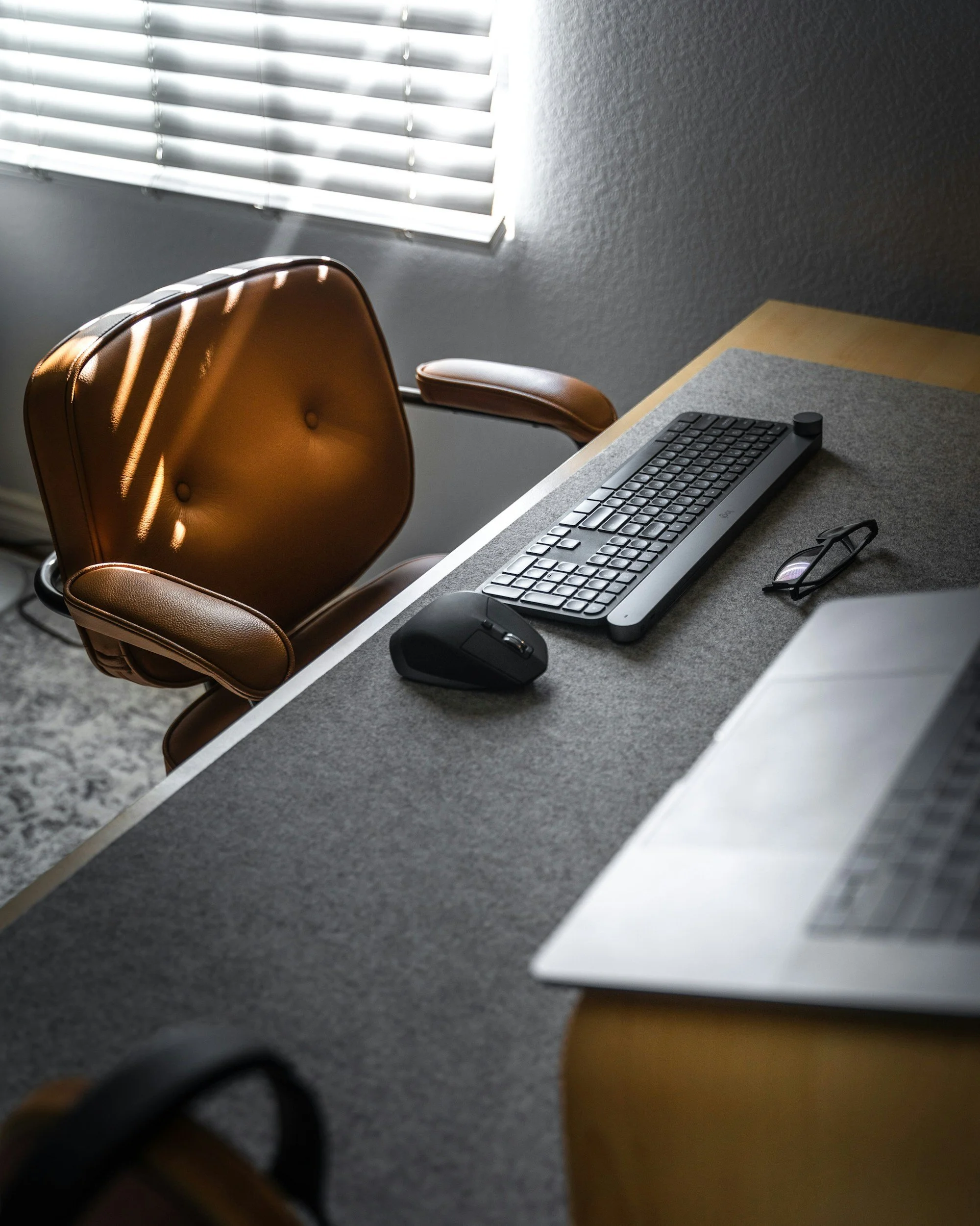 A brown leather office chair with wooden armrests next to a desk. On the desk, there's a computer keyboard, a computer mouse, and a pair of glasses. A window with horizontal blinds is on the wall, allowing sunlight to cast shadows.