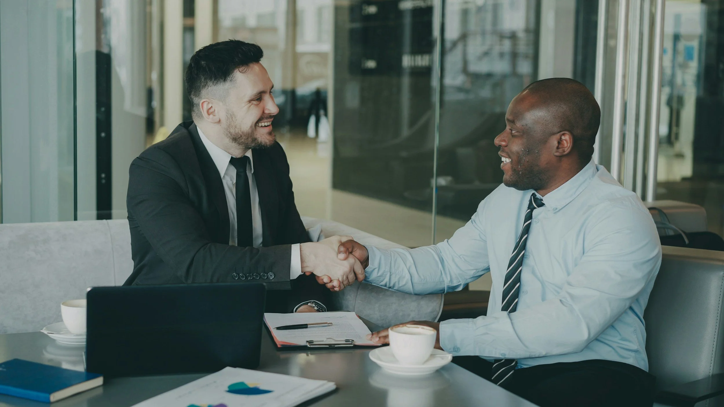Two men in business attire shaking hands and smiling in a modern office, with papers, a notebook, and coffee cups on the table.