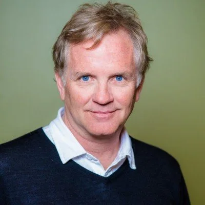 A portrait of a man with light skin, blond hair, blue eyes, wearing a white collared shirt and a dark sweater, standing against a green background.
