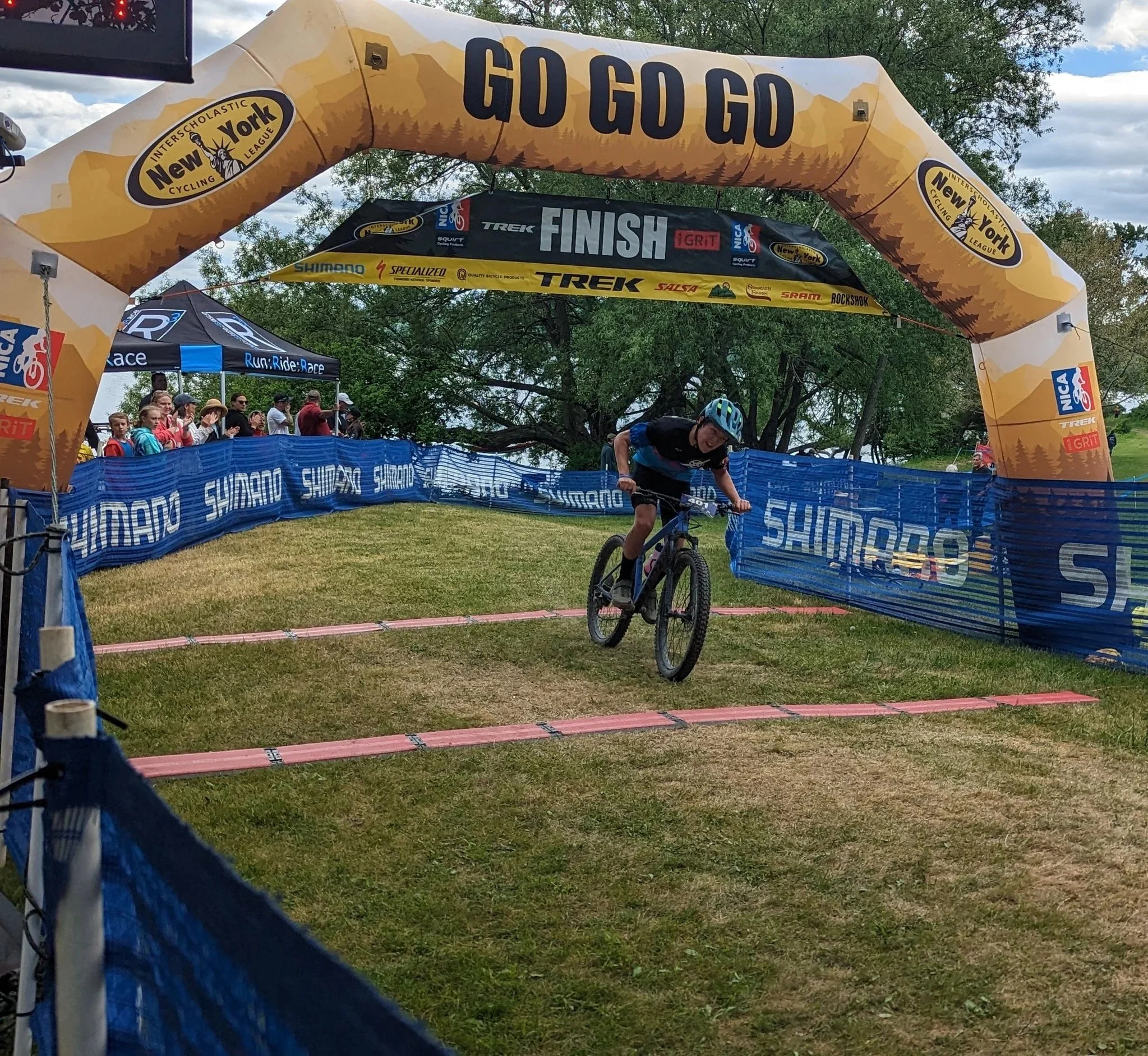 A cyclist jumping over a ramp during a race at a mountain biking event with spectators in the background, banners, and an archway reading 'GO GO GO'.