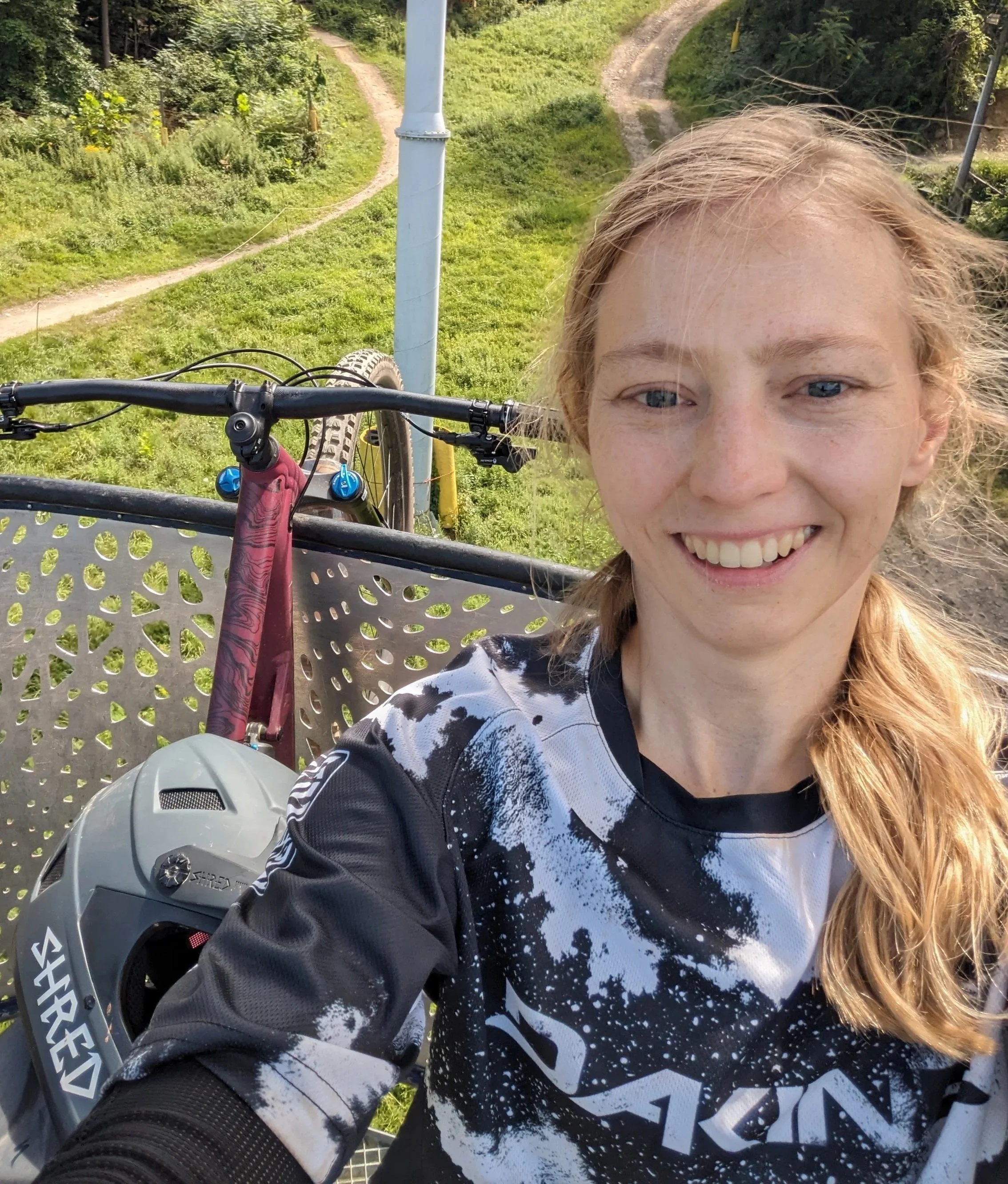 A smiling woman taking a selfie outdoors on a mountain biking trail, with her helmet and mountain bike visible in the background.