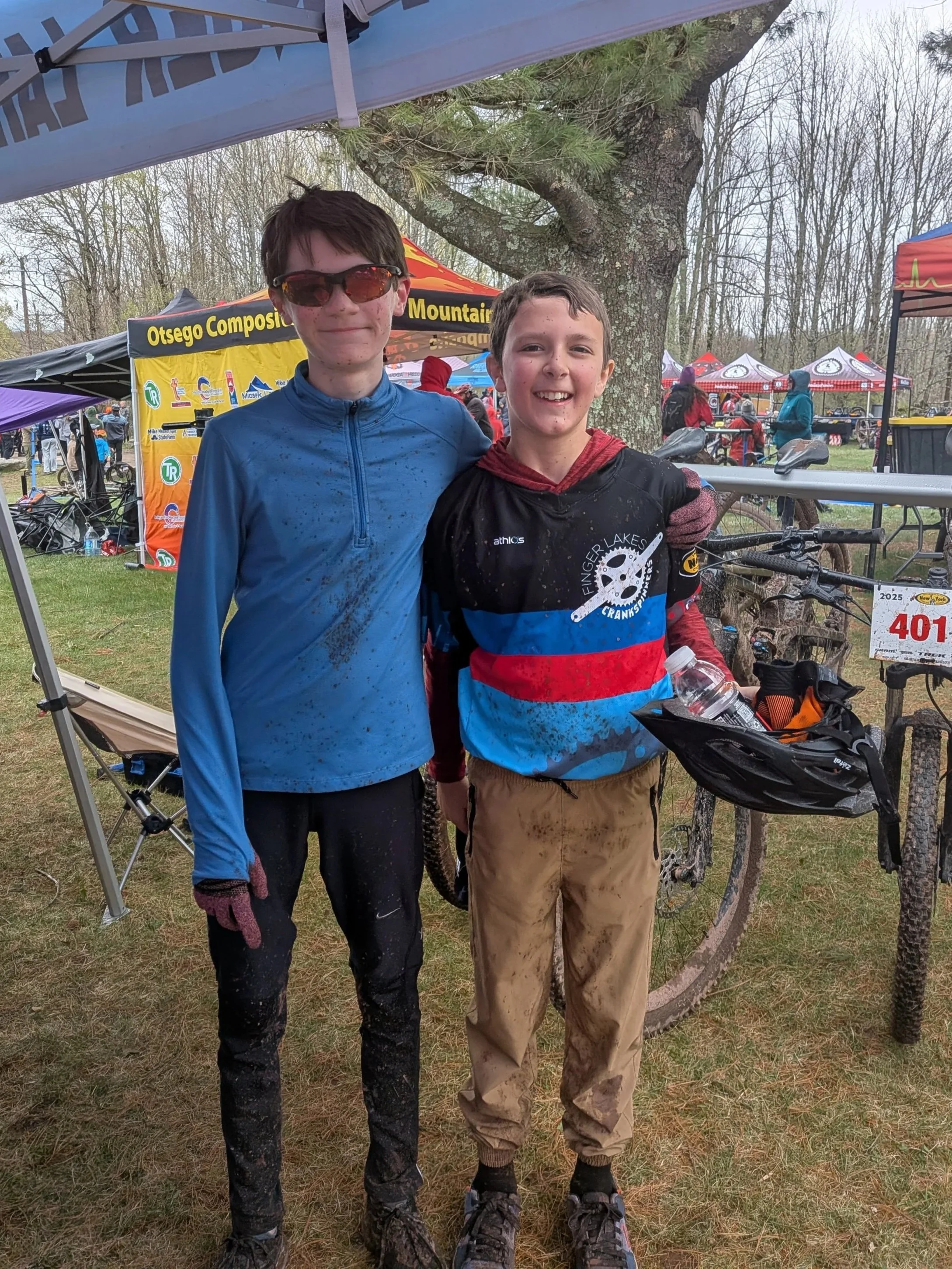 Two young boys standing outdoors after a trail race, smiling, with muddy clothing and a tent in the background.
