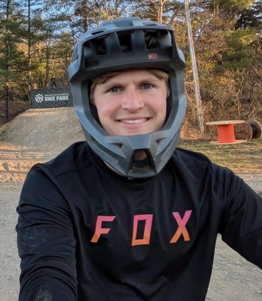 A young man wearing a black Fox shirt and a full-face motorcycle helmet, taking a selfie at a bike park with dirt jumps and trees in the background.