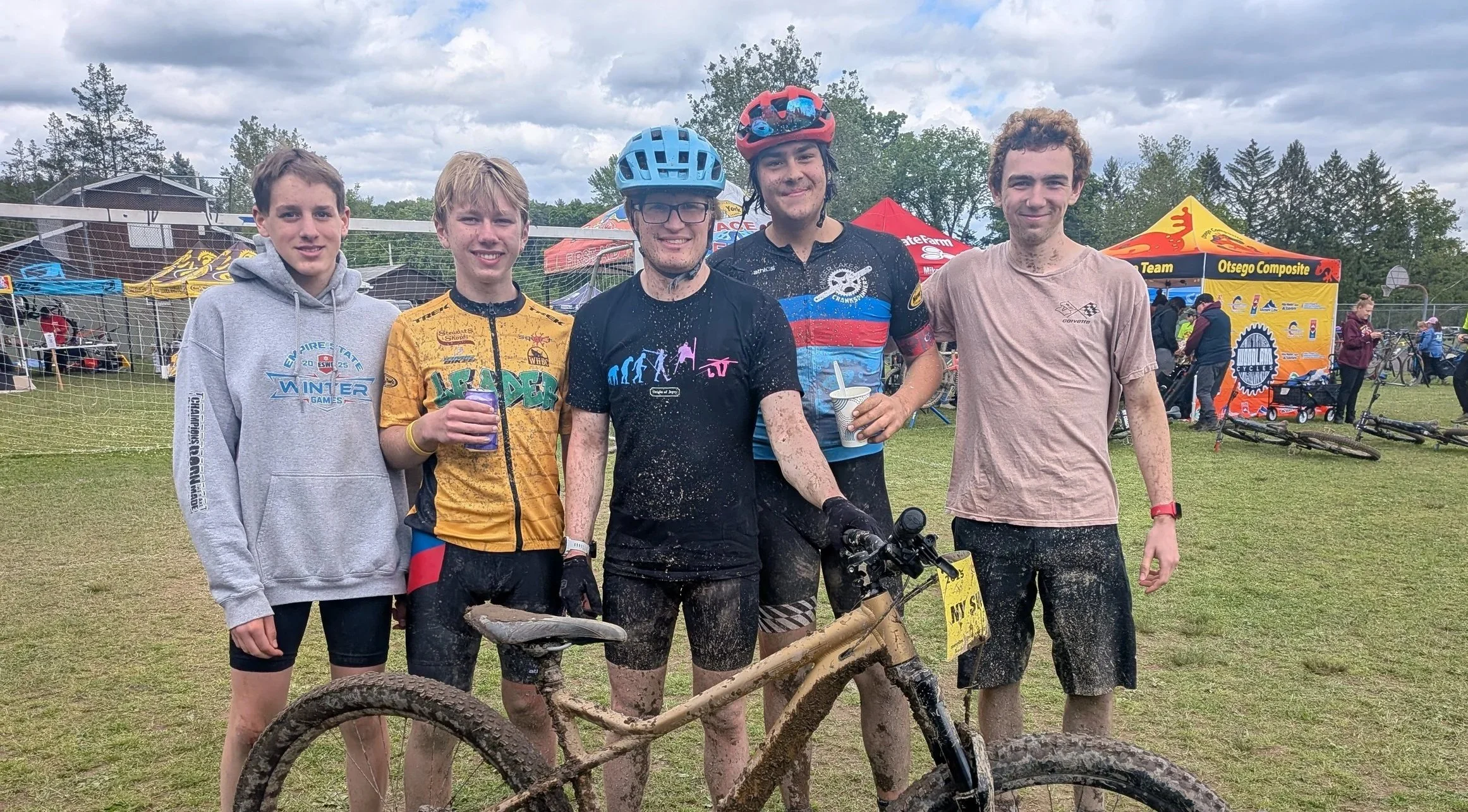 Six young men standing outdoors after a mountain biking event, some with mud on their clothes and bikes, with tents and people in the background.