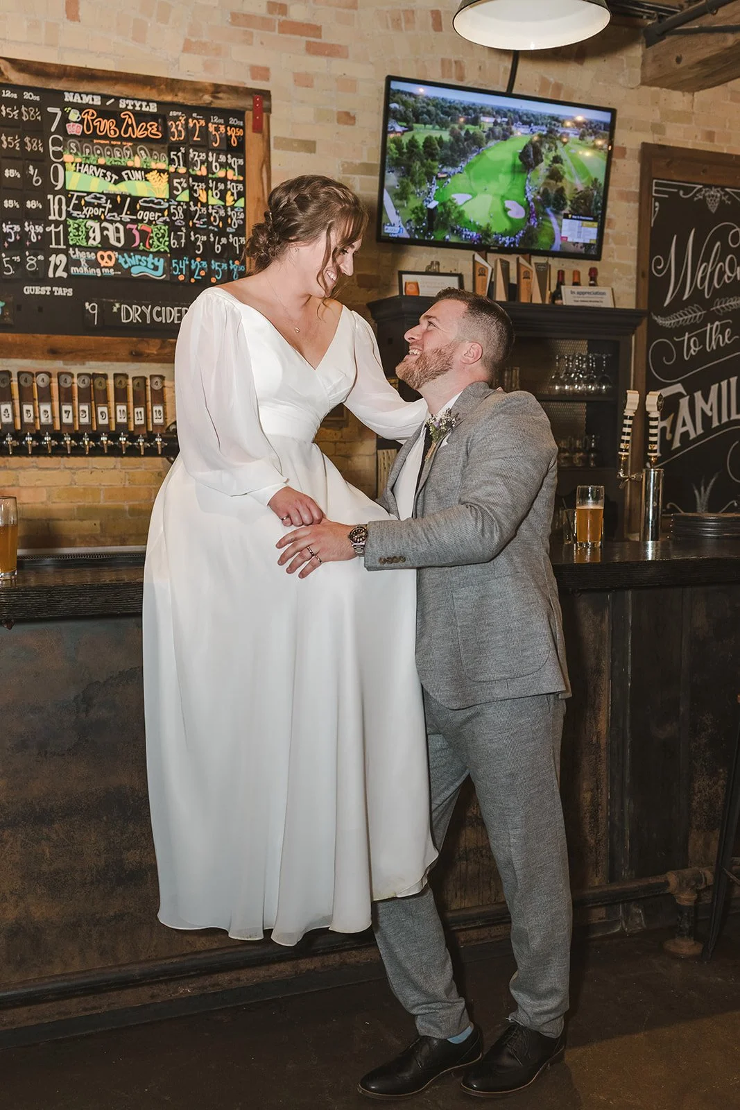 Bride Sitting on Bar Top in Wedding Dress with Groom Embracing Her Fedora Media Cambridge Ontario Four Fathers Brewery.jpg