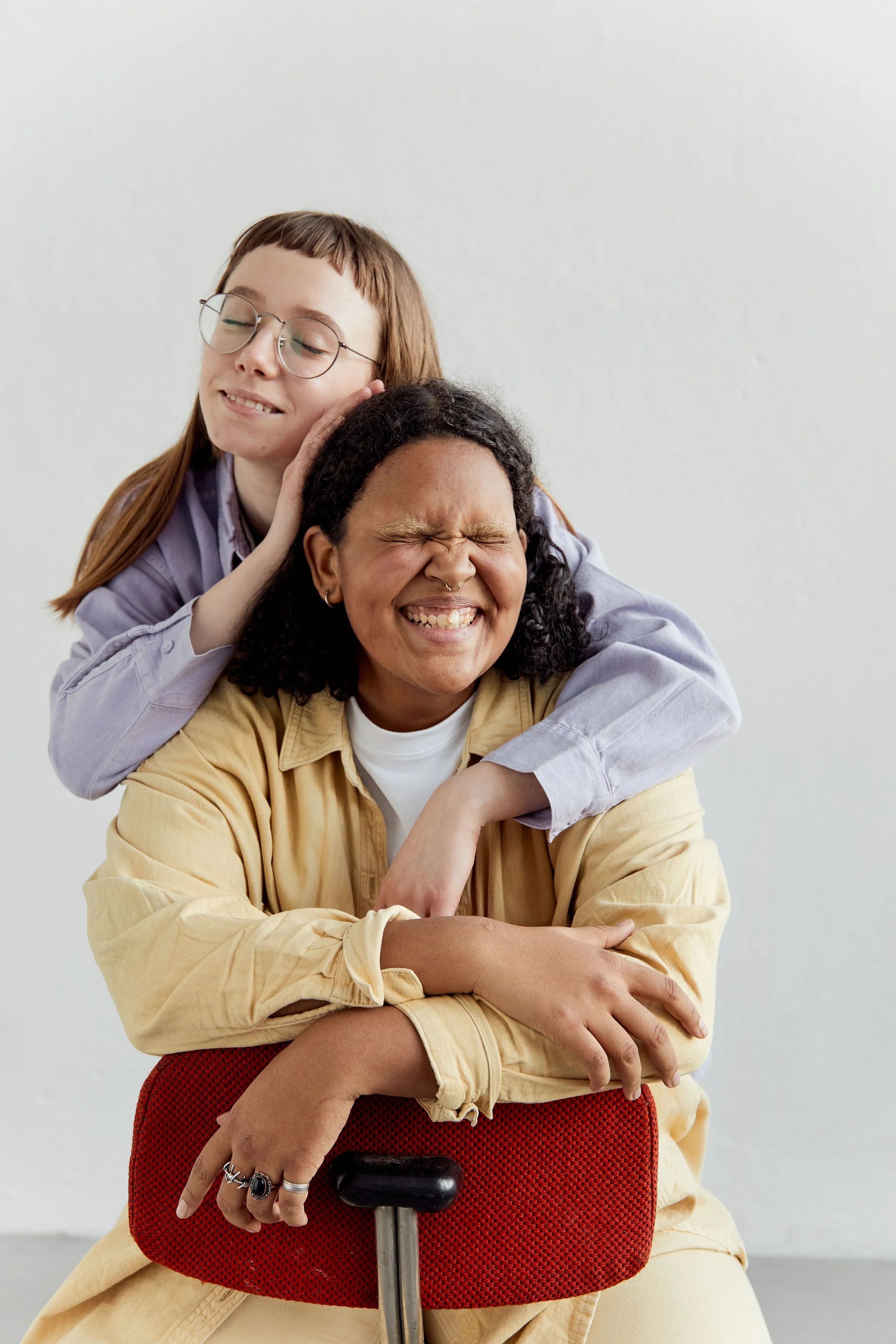Two queer people sharing a warm, laughing hug and genuine connection, one wearing glasses