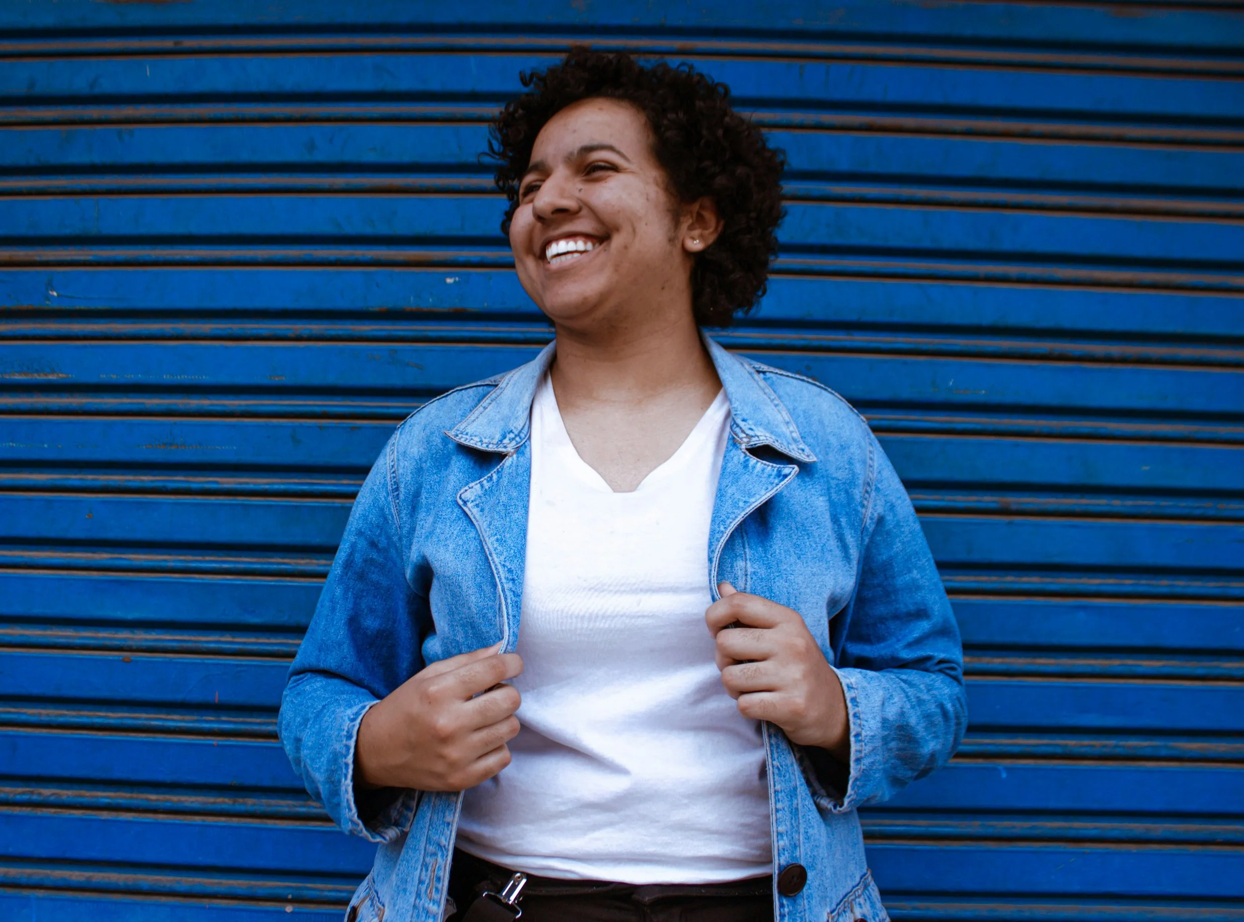 Person in a denim jacket smiling confidently against a blue corrugated wall