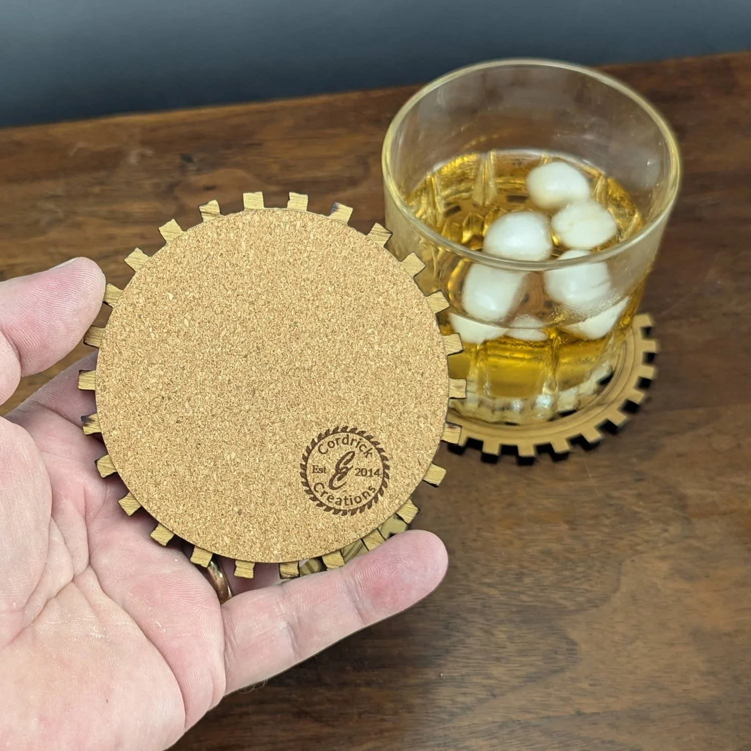 Back view of The Operator wooden coaster showing natural cork backing with engraved Cordrick Creations logo, whiskey glass behind.
