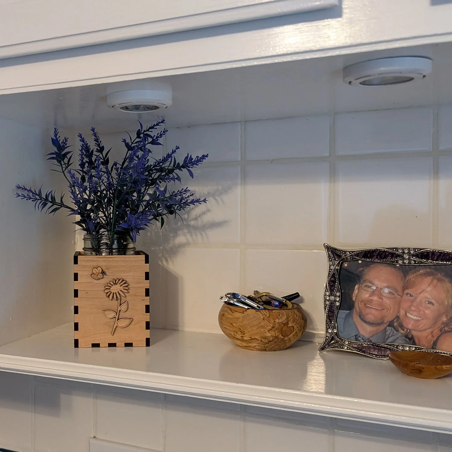 Maple Bloom & Buzz vase box with purple flowers displayed on white kitchen shelf beside wooden bowl and photo frame.