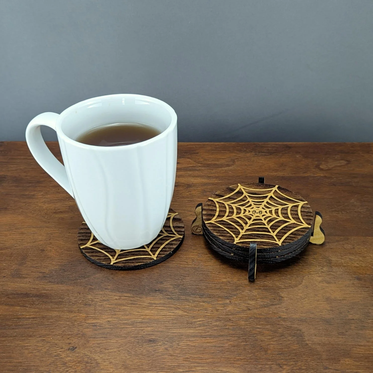 Tea mug resting on a spiderweb coaster beside a stacked set in a black wooden holder, all displayed on a wooden tabletop.