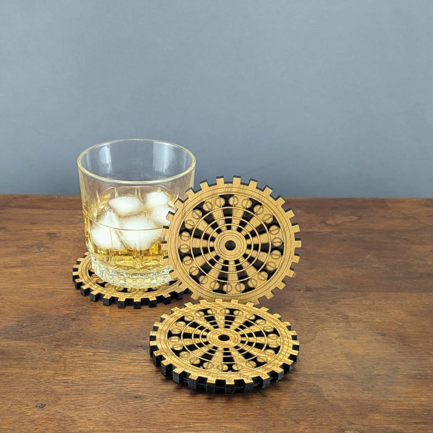 Glass of whiskey with ice placed on a wooden gear coaster, additional coasters arranged beside it.