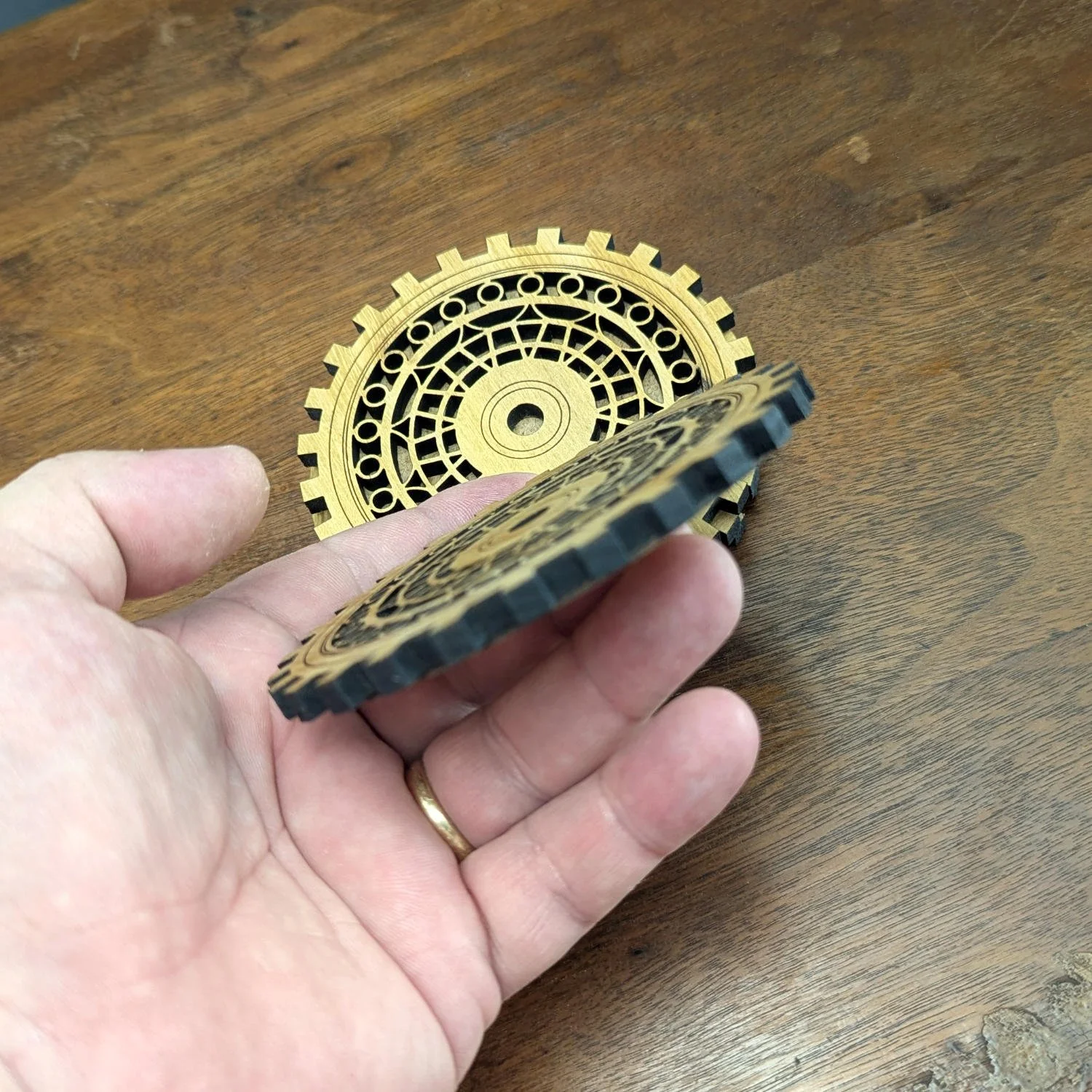 Hand holding The Precisionist wooden coaster, showing gear-shaped edge and stacked coasters in the background.