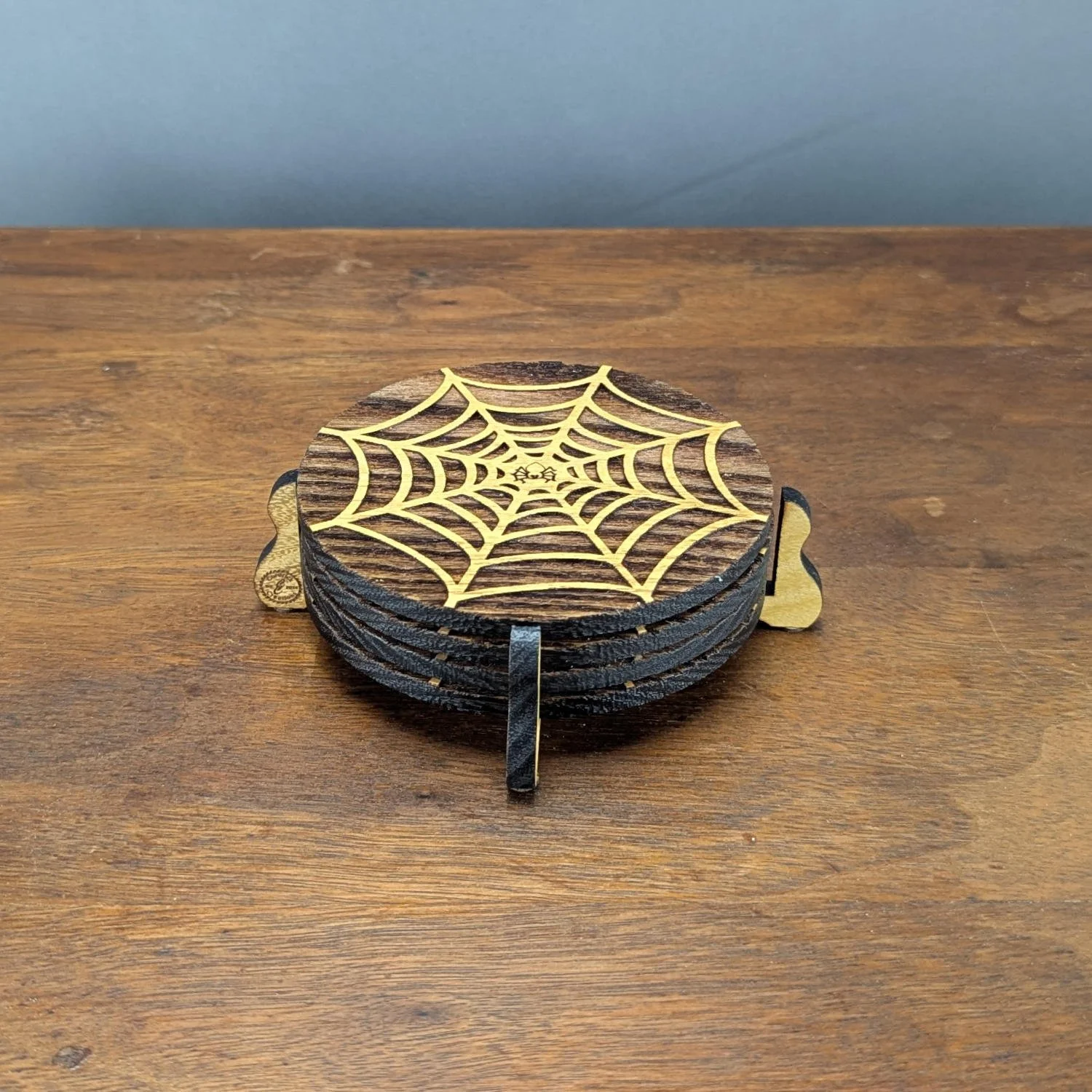 Angled view of four spiderweb wooden coasters stacked together in a holder, showing the engraved golden web pattern.