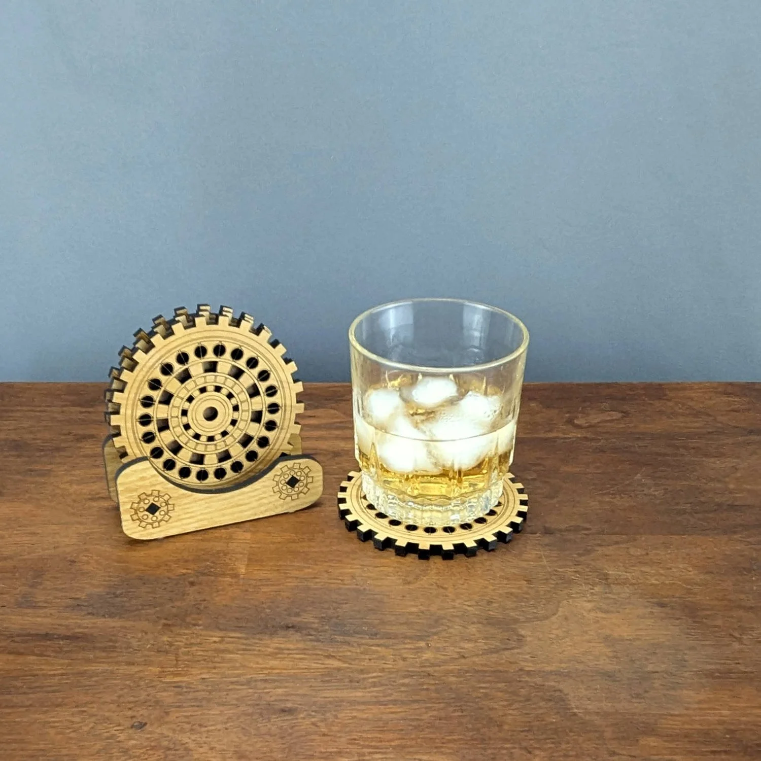 Wooden gear coasters displayed in a holder with gear designs, alongside a glass of whiskey resting on another coaster.