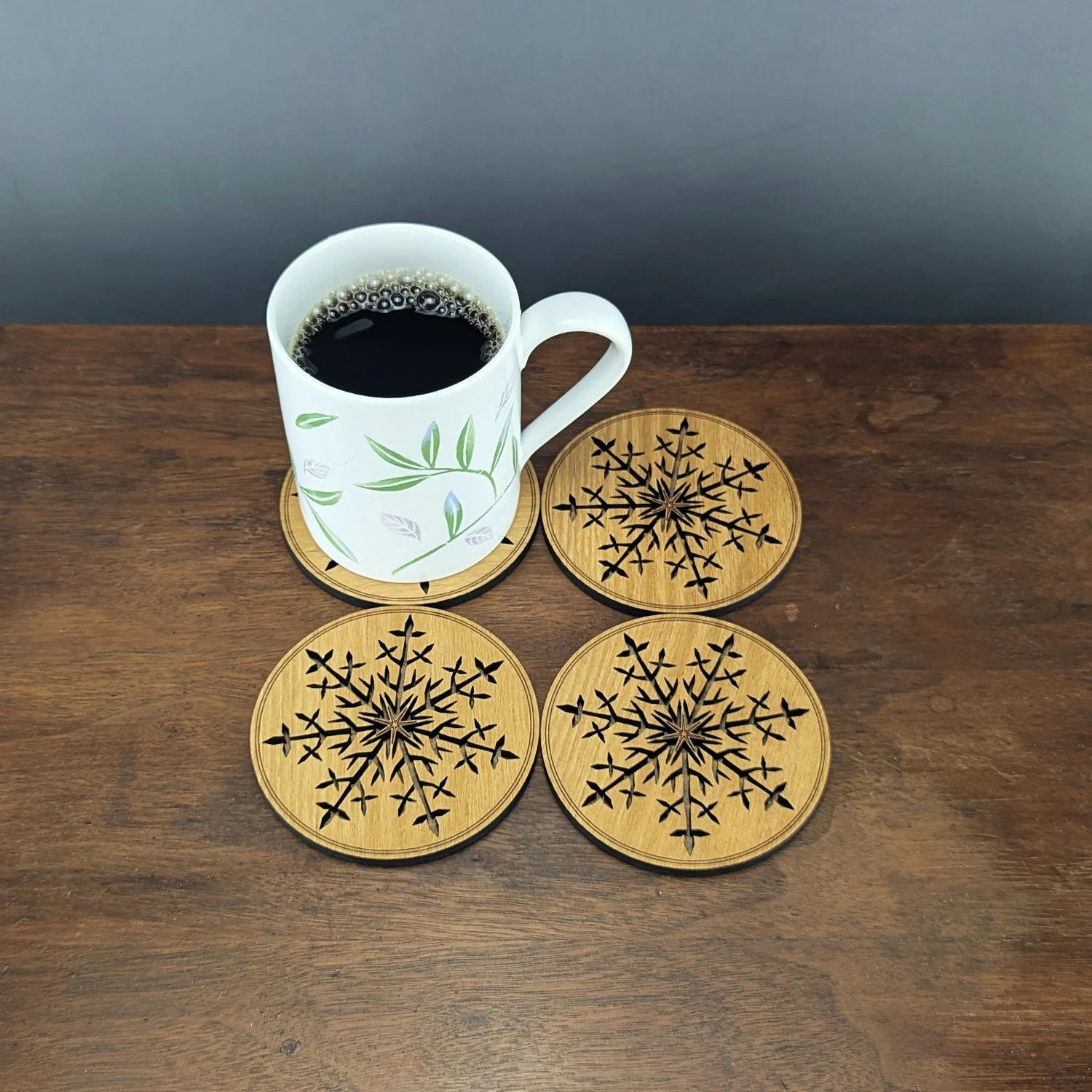 Overhead view of four Black Locust snowflake coasters arranged around a coffee mug on a wooden table.