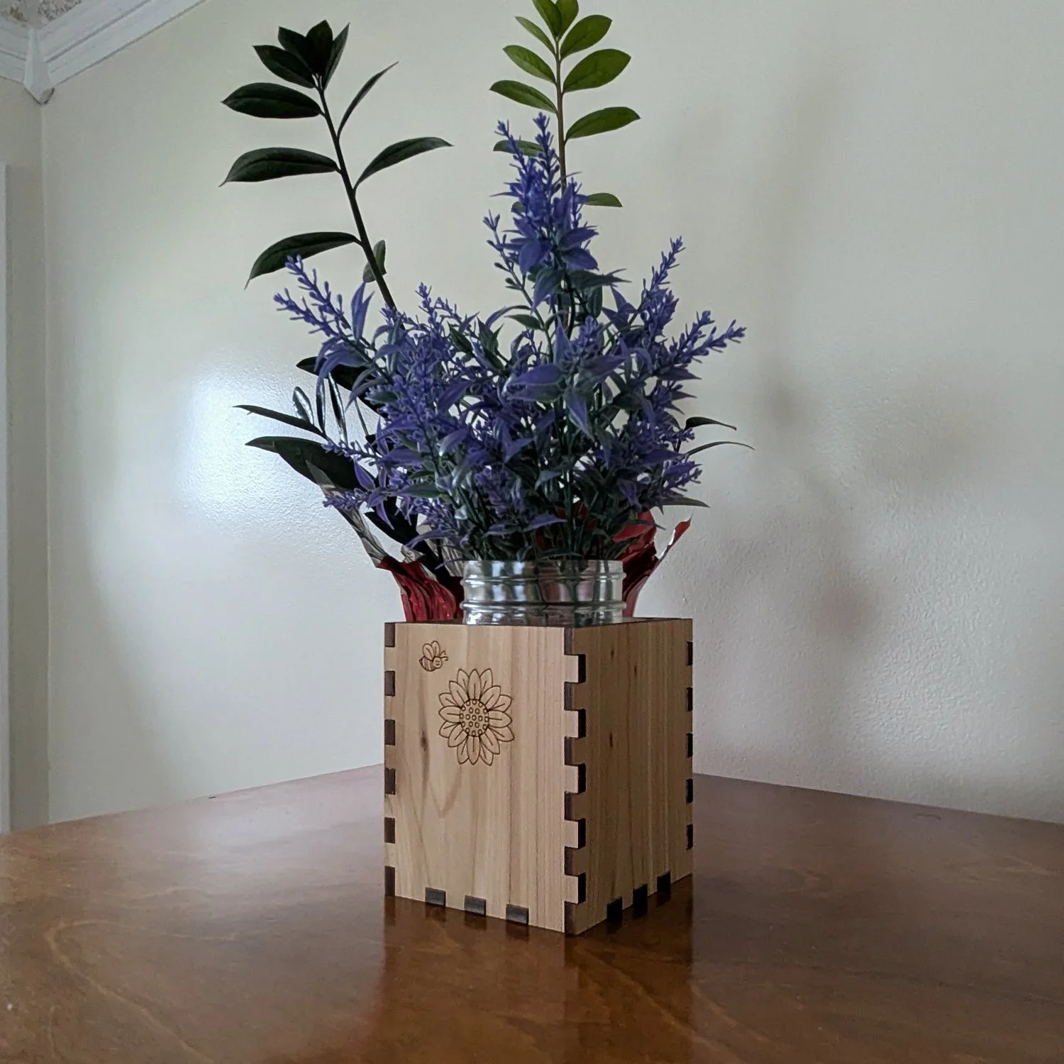Angled view of the bee-and-sunflower engraved wooden vase sleeve with purple flowers in a mason jar, displayed on a wood table.