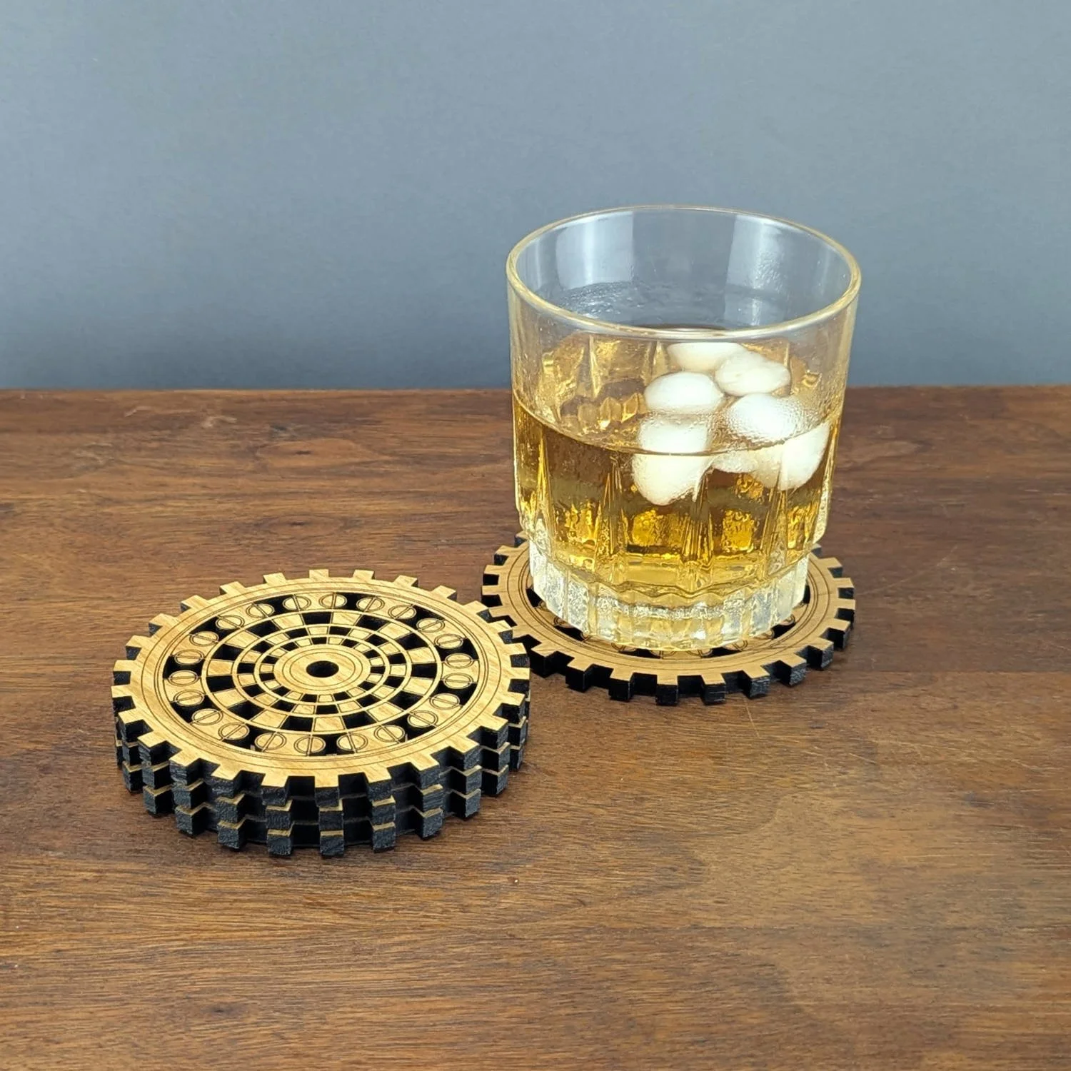 Glass of whiskey with ice placed on The Operator wooden gear coaster, second coaster stacked beside it on wooden surface.