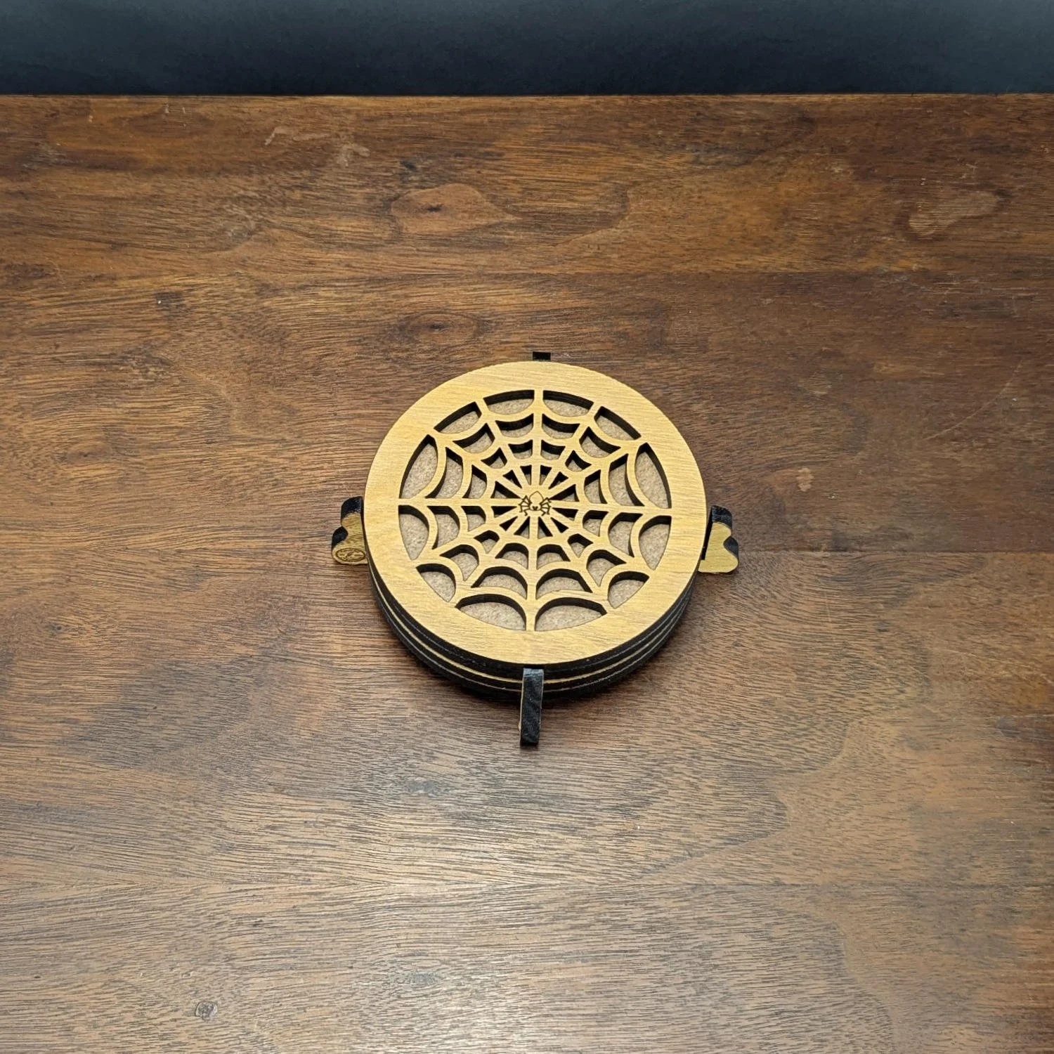 Stack of Iron Web wooden drink coasters in spiderweb pattern, neatly arranged in a holder on a wood surface