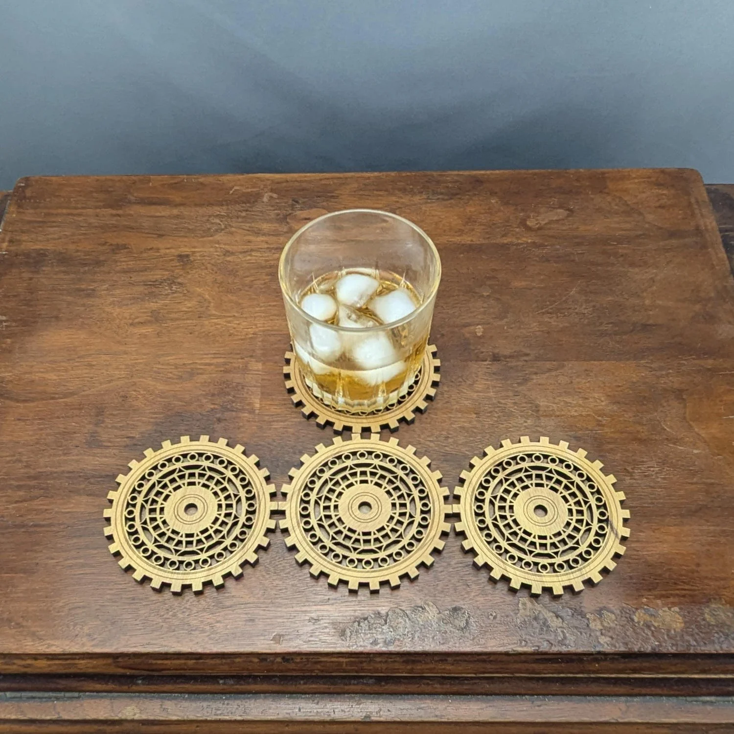 Set of four Precisionist wooden gearwork coasters displayed on a table, one holding a whiskey glass with ice while the others are lined up neatly.