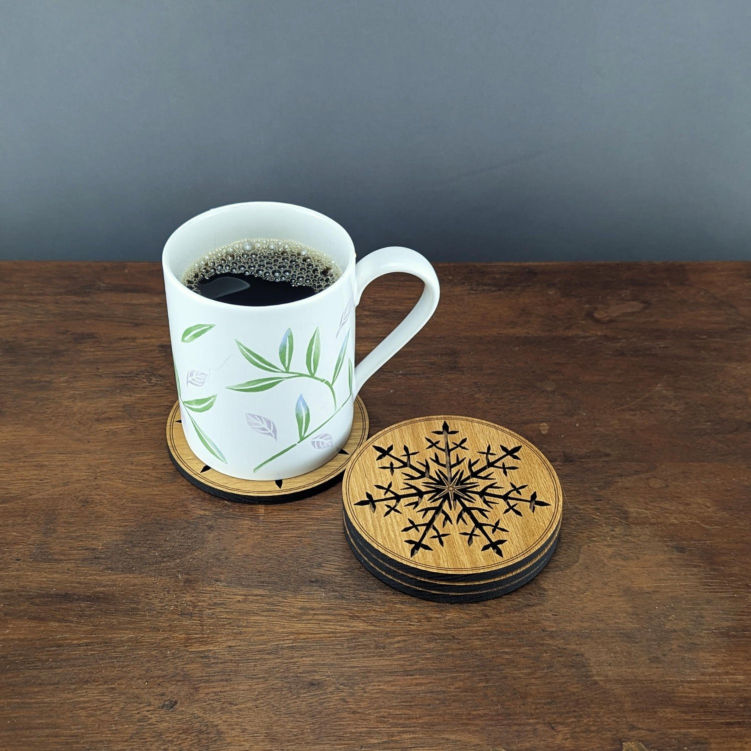 White coffee mug sitting on a Black Locust snowflake coaster with a stacked set of laser-cut wooden snowflake coasters beside it on a dark wood table.