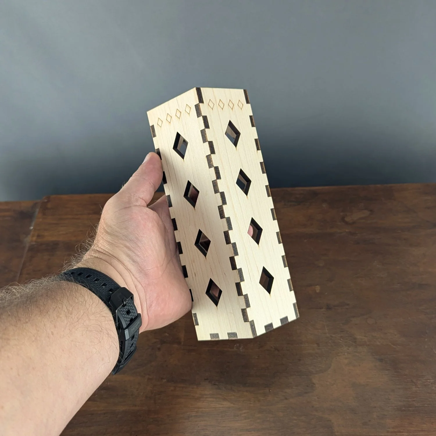 Hand holding maple wood vase sleeve to show size and detail of diamond cutouts and laser-cut finger joints.