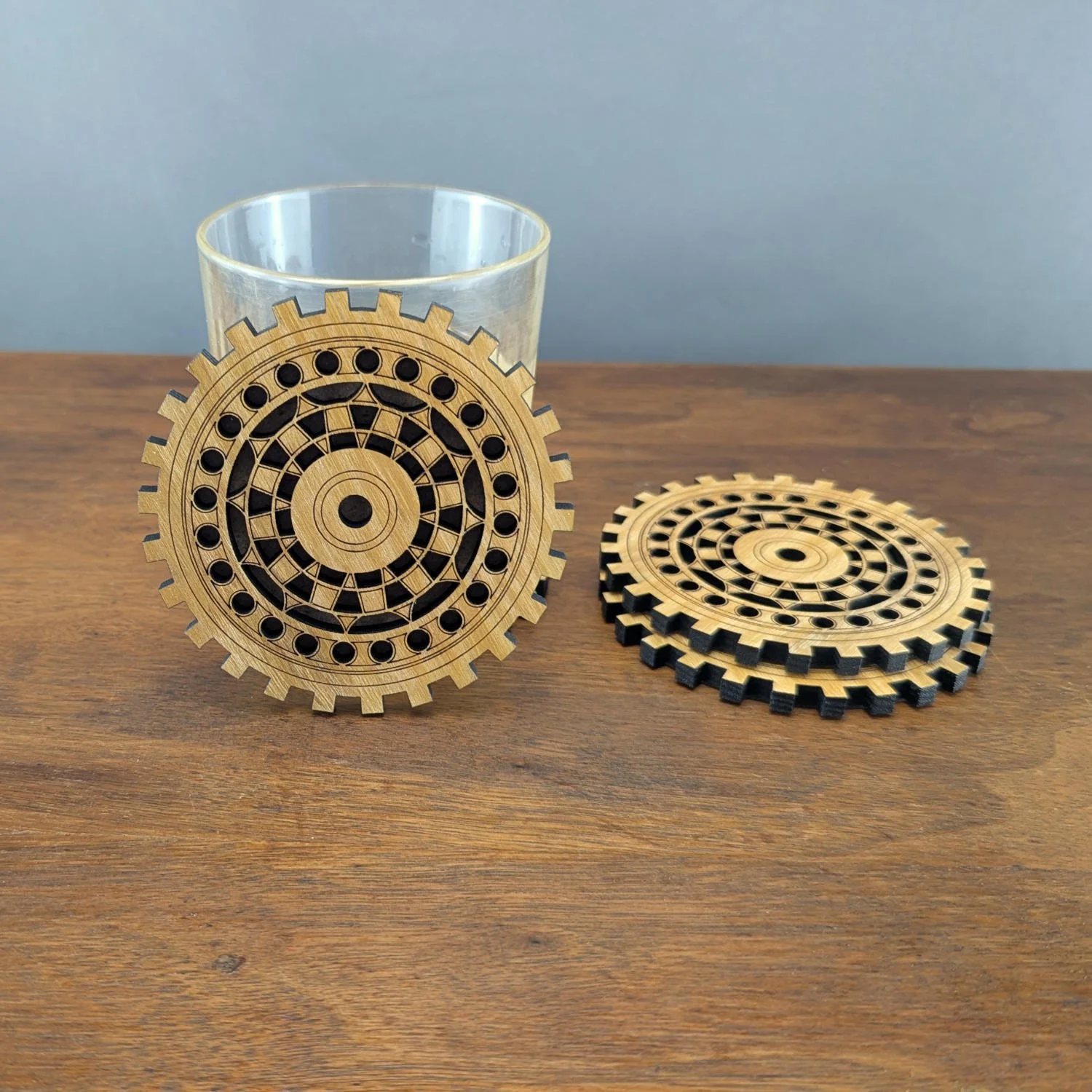 Close-up of The Engineer wooden gear coaster propped against a glass with another coaster flat on table.
