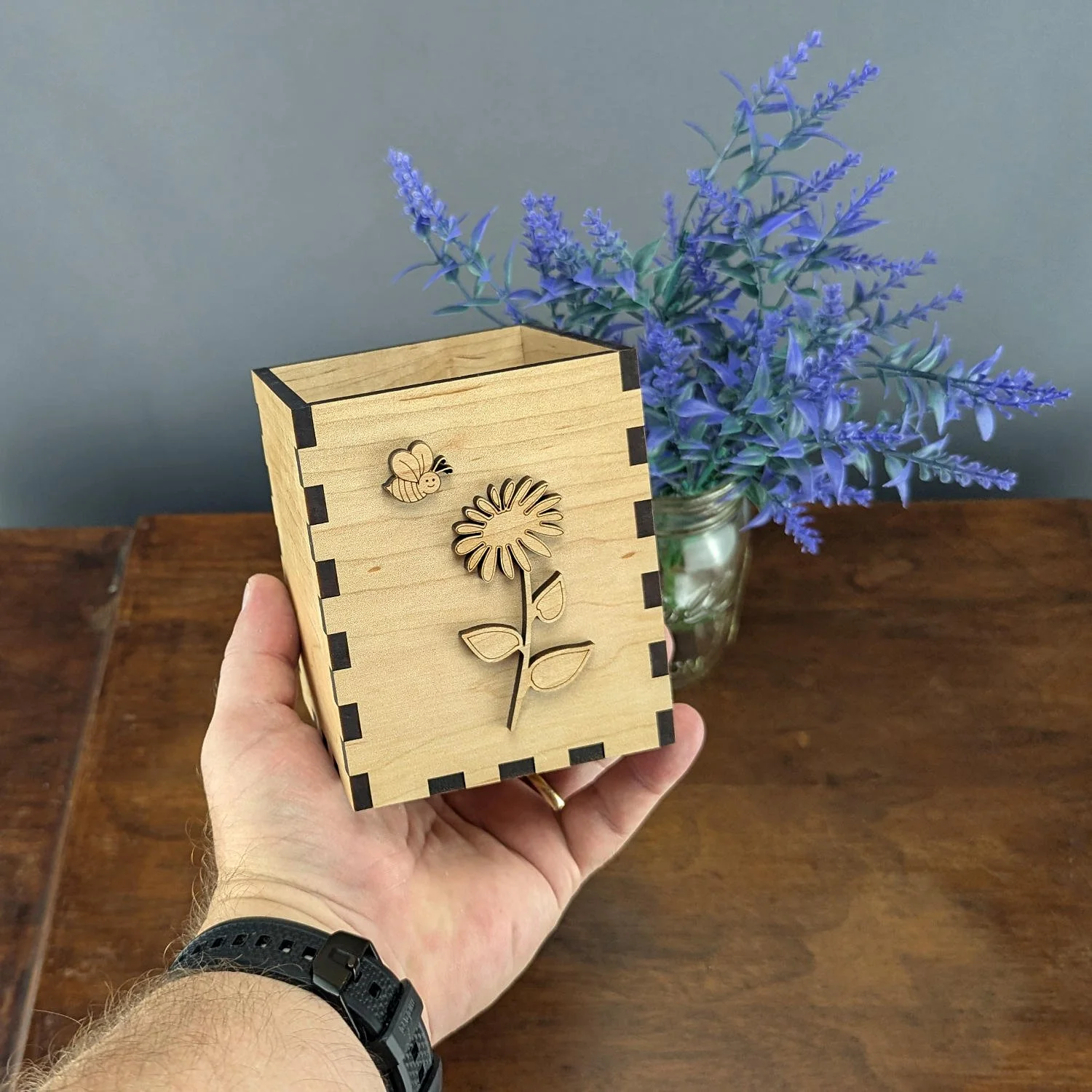 Hand holding the maple Bloom & Buzz vase box in front of purple flowers, showing scale and craftsmanship.
