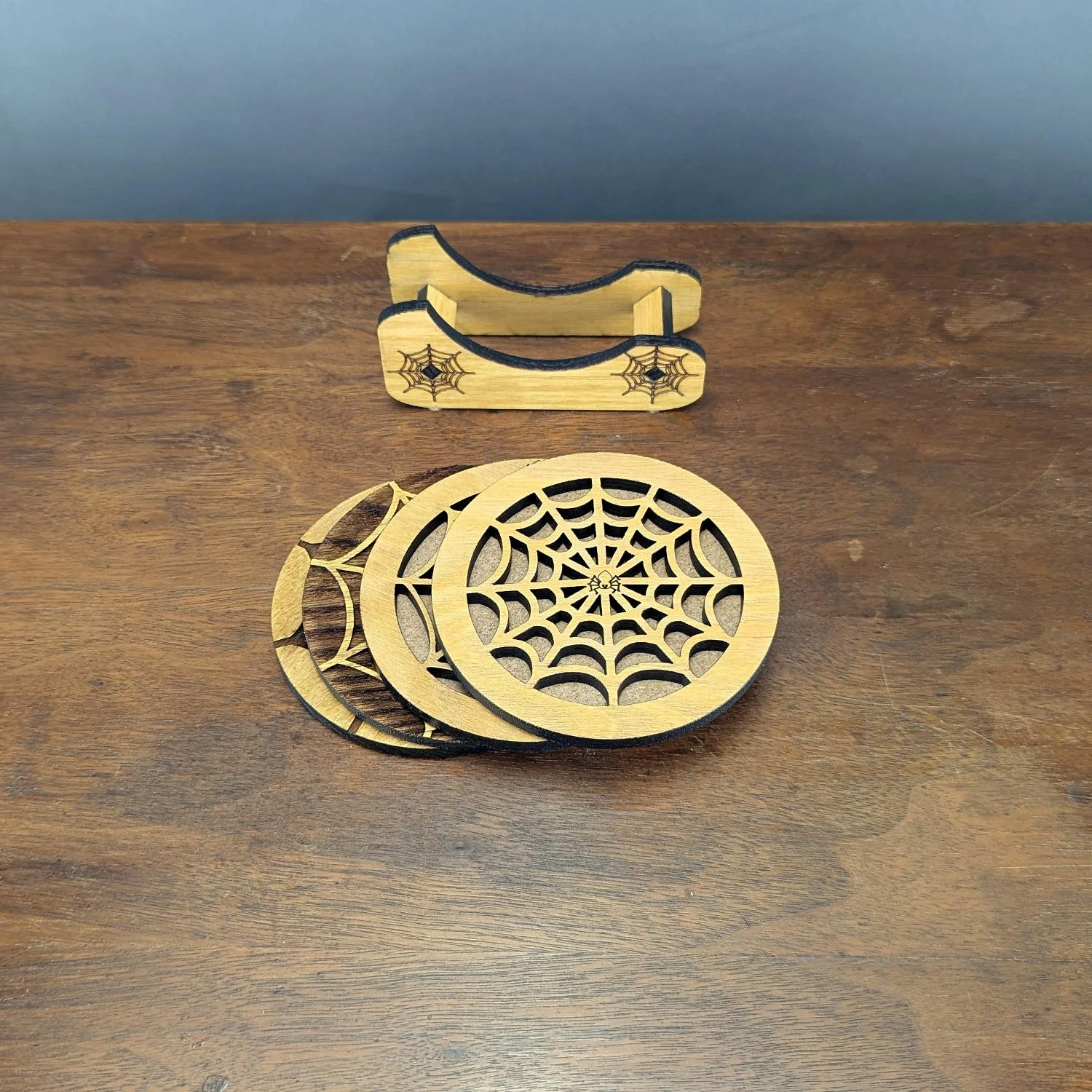 Stack of laser-cut spiderweb coasters beside matching engraved holder, cork-backed and finished with matte acrylic.