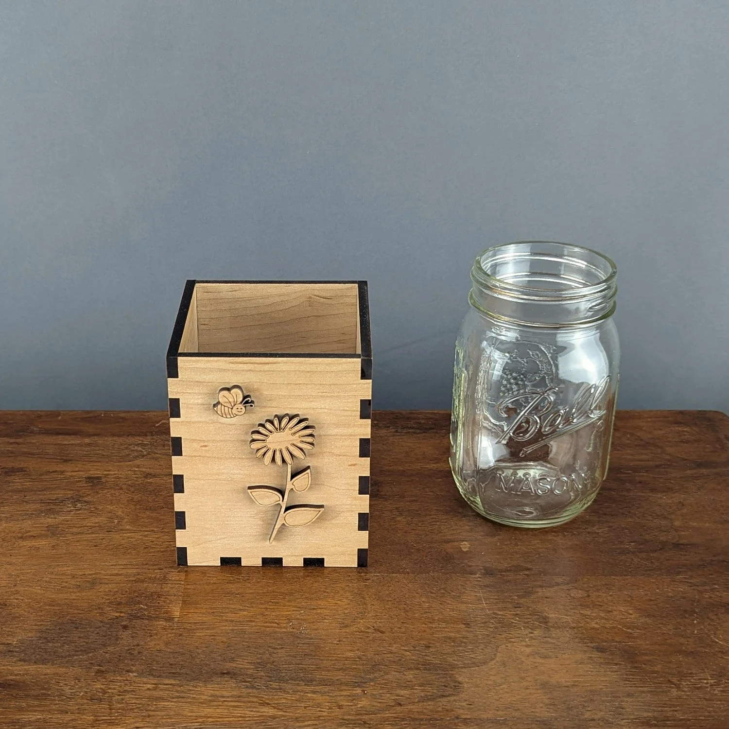Handmade maple wood vase sleeve with laser-cut bee and flower design beside clear mason jar on wooden table.