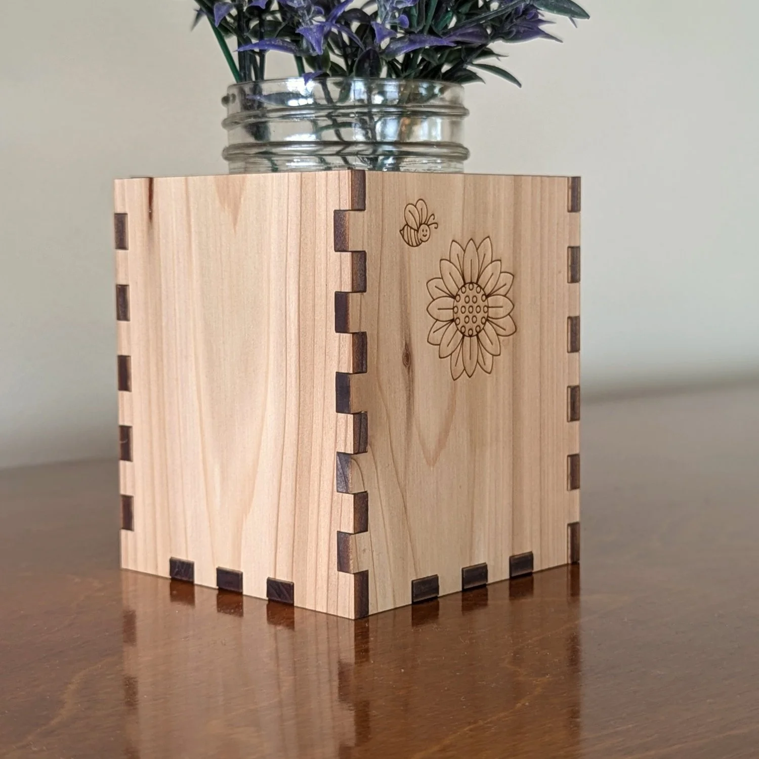 Side-angle photo of the wooden mason jar vase sleeve with engraved bee and sunflower, holding purple flowers in a glass mason jar.