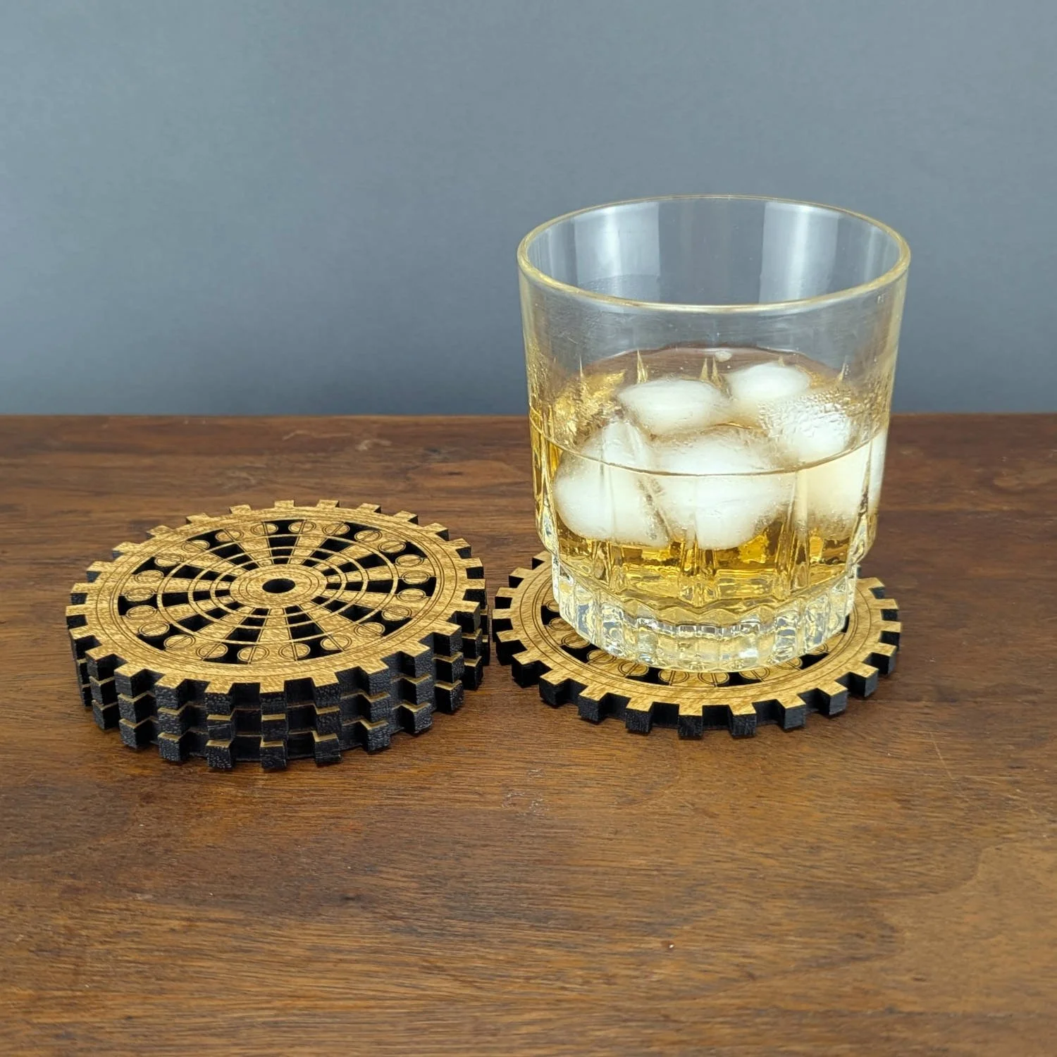 Stack of wooden gear coasters next to a glass of whiskey on a coaster, displayed on a wooden surface.