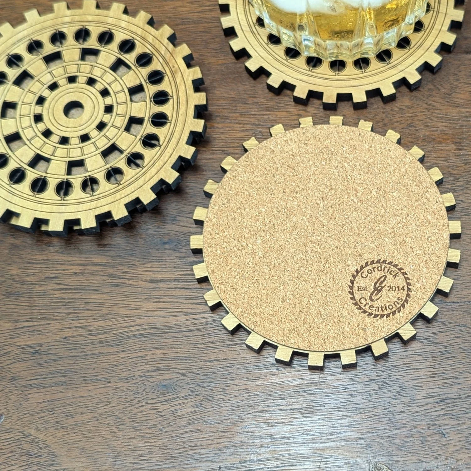 Back view of a cork-lined gear-shaped coaster with engraved maker’s mark, shown alongside other engraved coasters and a glass of whiskey.