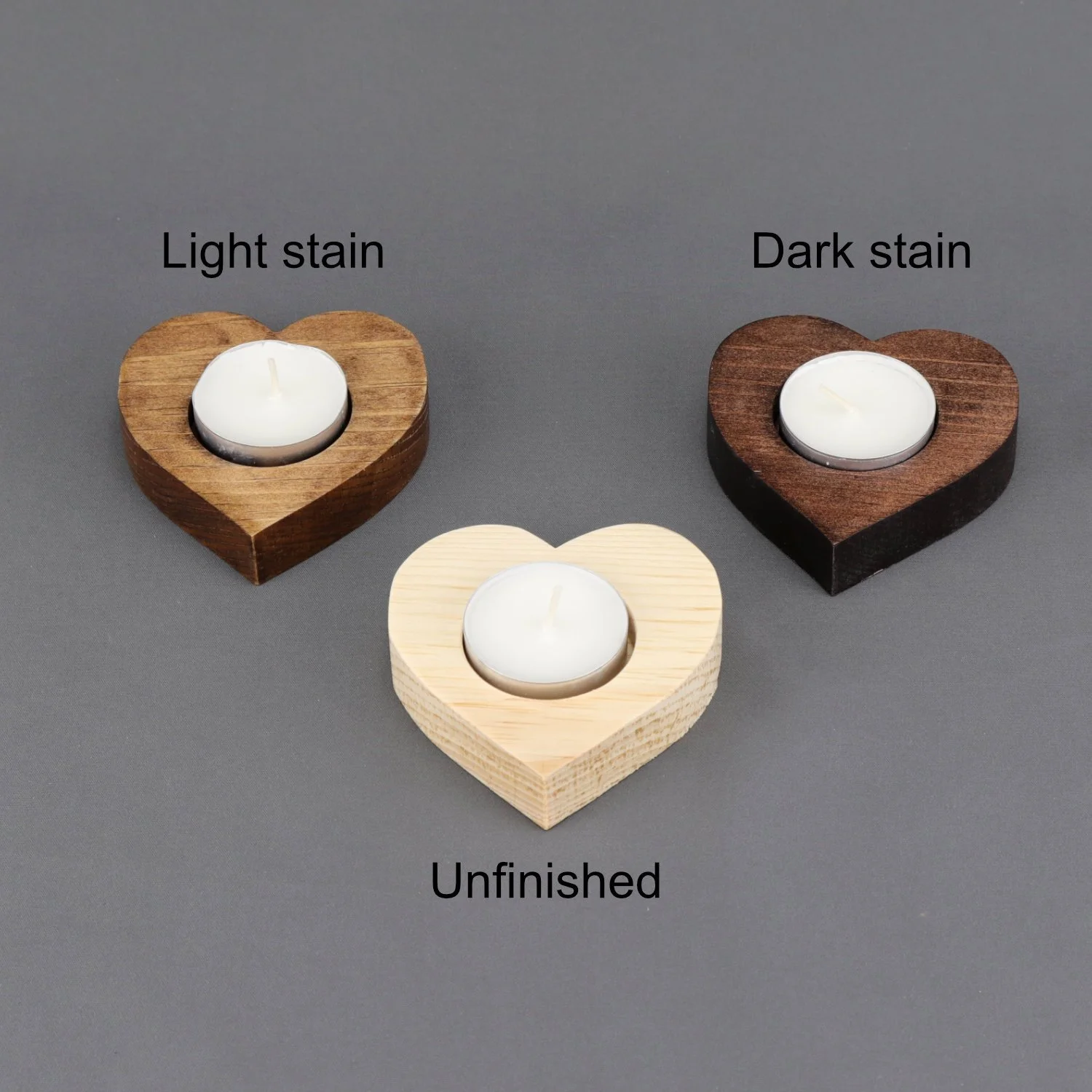 Three heart-shaped wooden candle holders, one unfinished light-colored, one dark brown, and one medium brown, with tea light candles, on a gray surface, labeled 'Light stain,' 'Dark stain,' and 'Unfinished.'