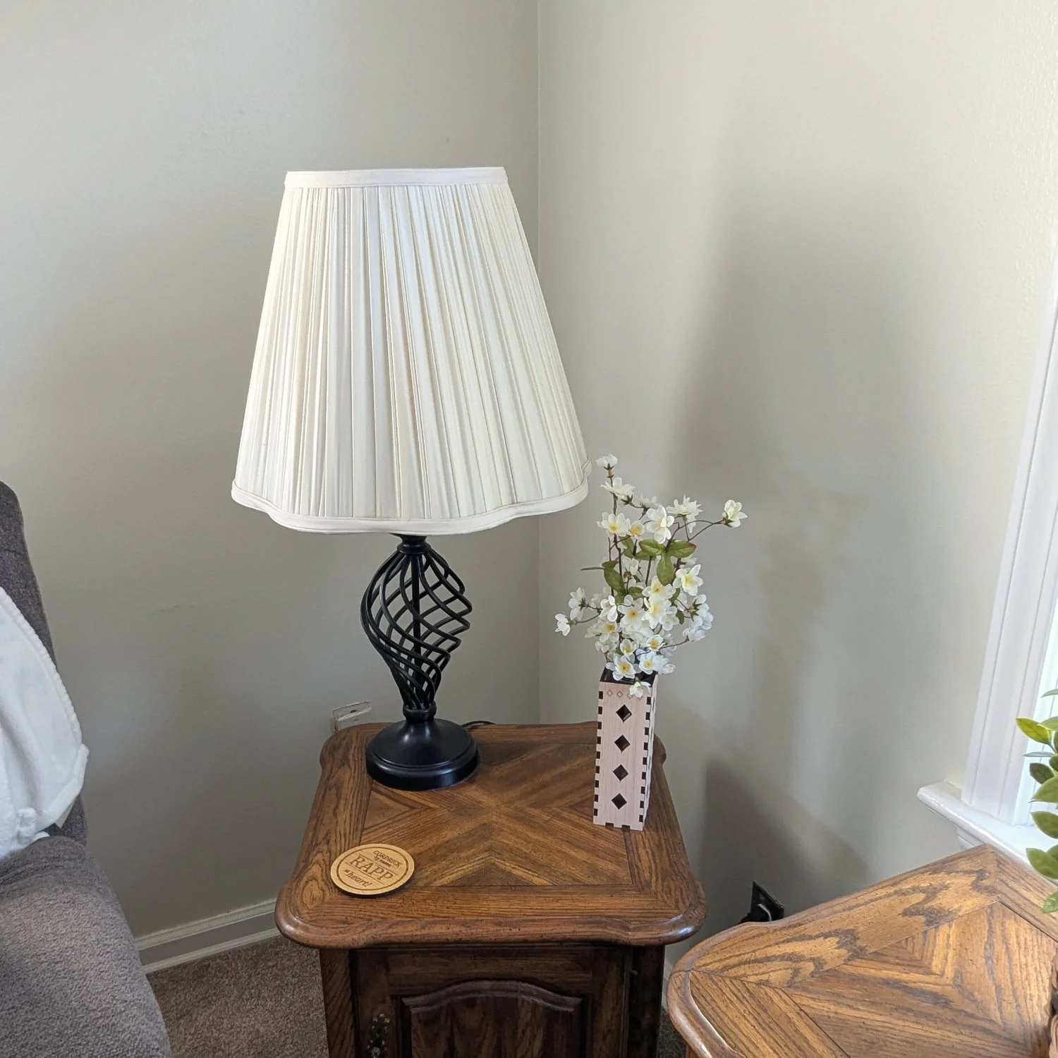 Maple wooden vase sleeve with diamond cutouts displayed on an end table beside a lamp, holding white flowers.