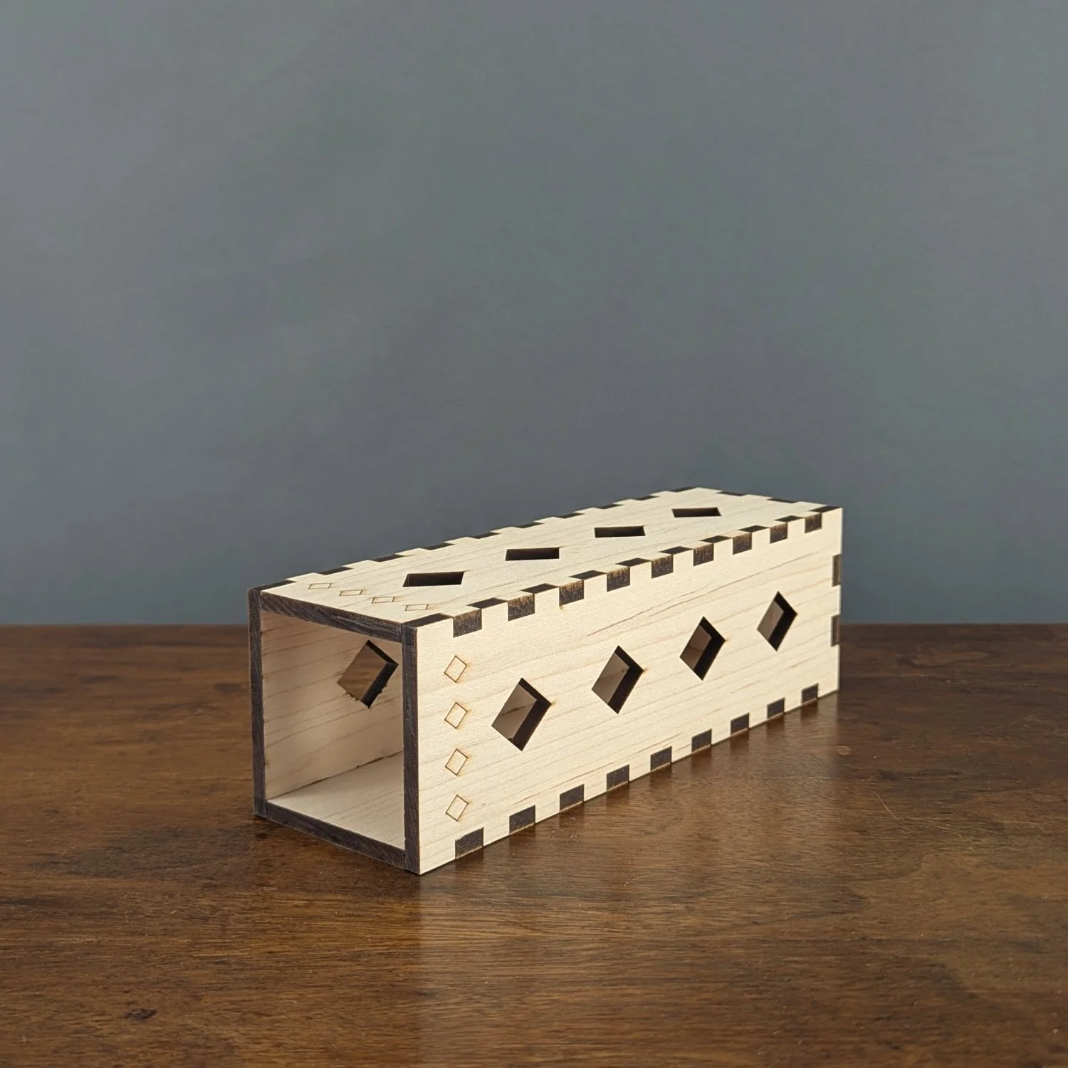 Handmade maple wooden vase sleeve with diamond cutout design, shown laid on its side to display finger-joint construction and laser-cut details.