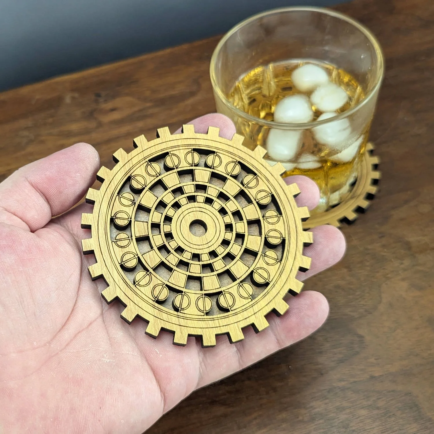 Hand holding The Operator wooden gear coaster above a table with a glass of whiskey in the background.