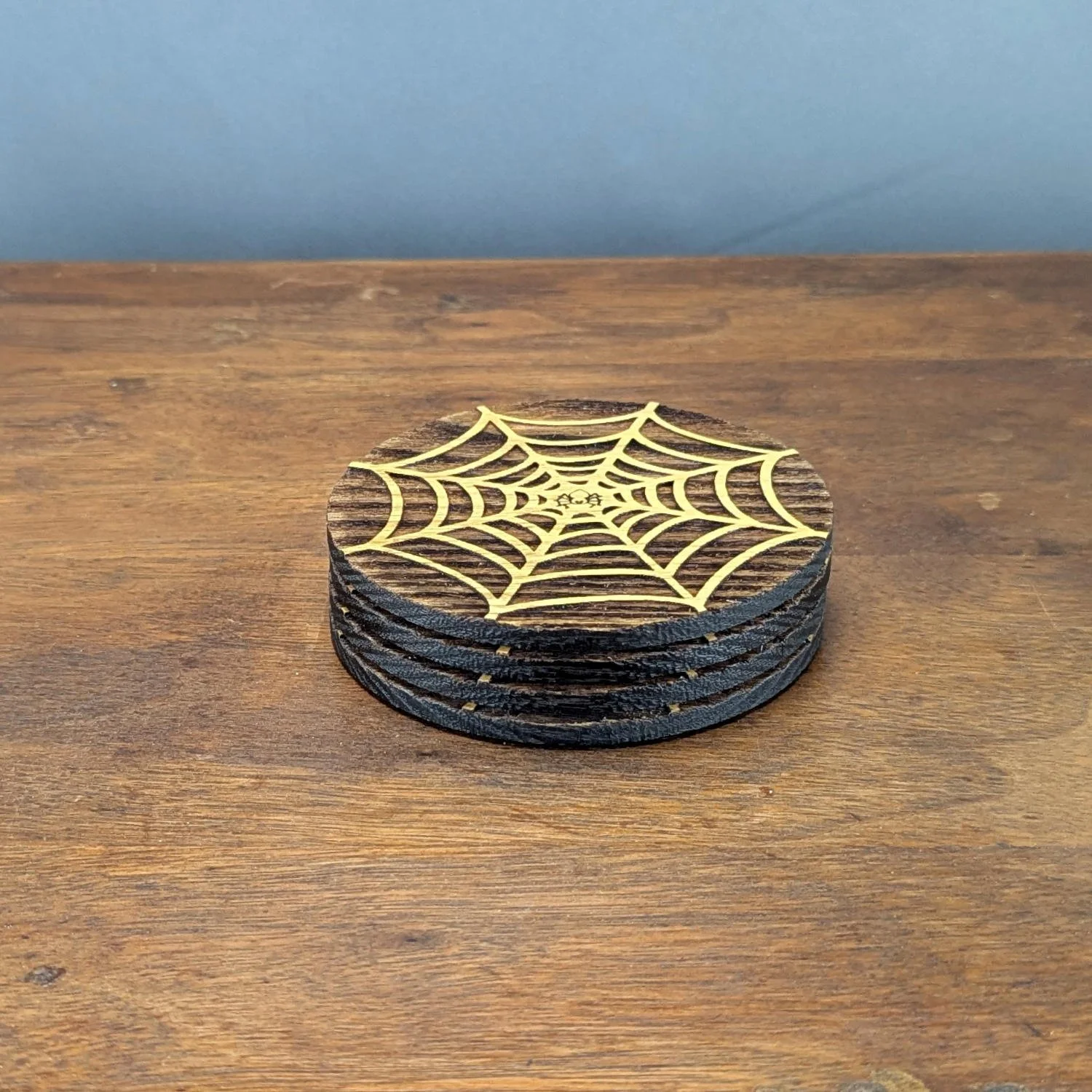 Side view of a neatly stacked set of four spiderweb wooden coasters on a wooden surface, without a holder.