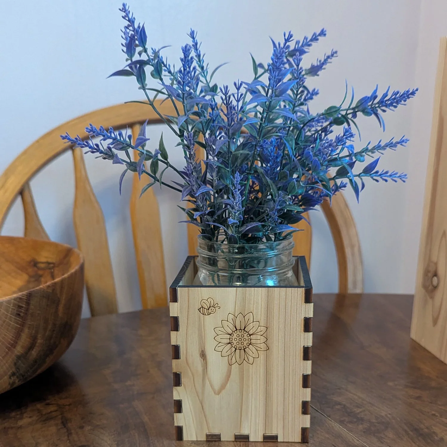 Wooden bee-and-sunflower engraved vase sleeve with mason jar and purple flowers, displayed on a dining table with wooden chairs in the background.