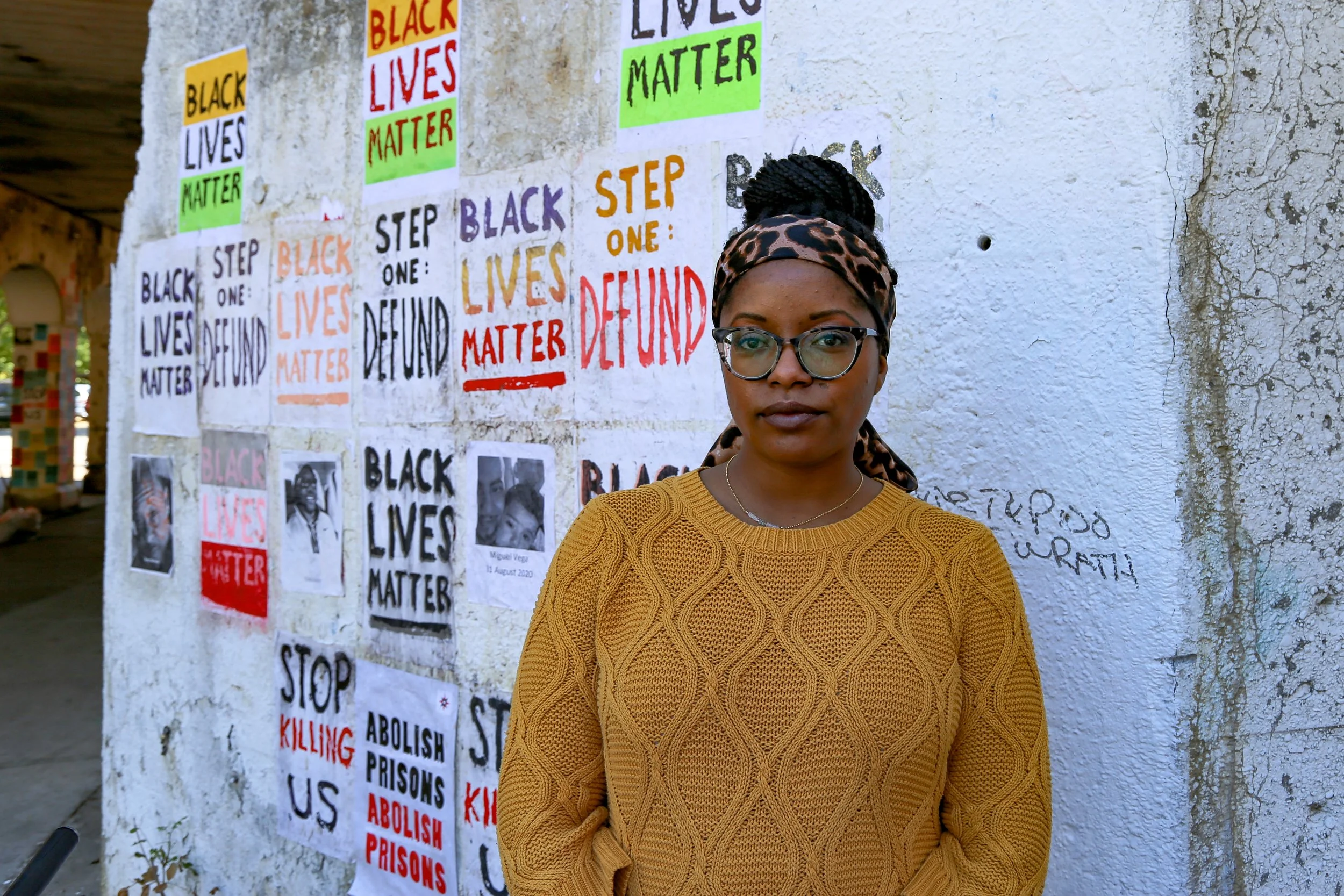 A woman wearing glasses, a leopard print headscarf, and a mustard yellow sweater standing in front of a white wall covered with posters and signs with messages like "Black Lives Matter," "Stop Killing Us," and "Abolish Prisons."