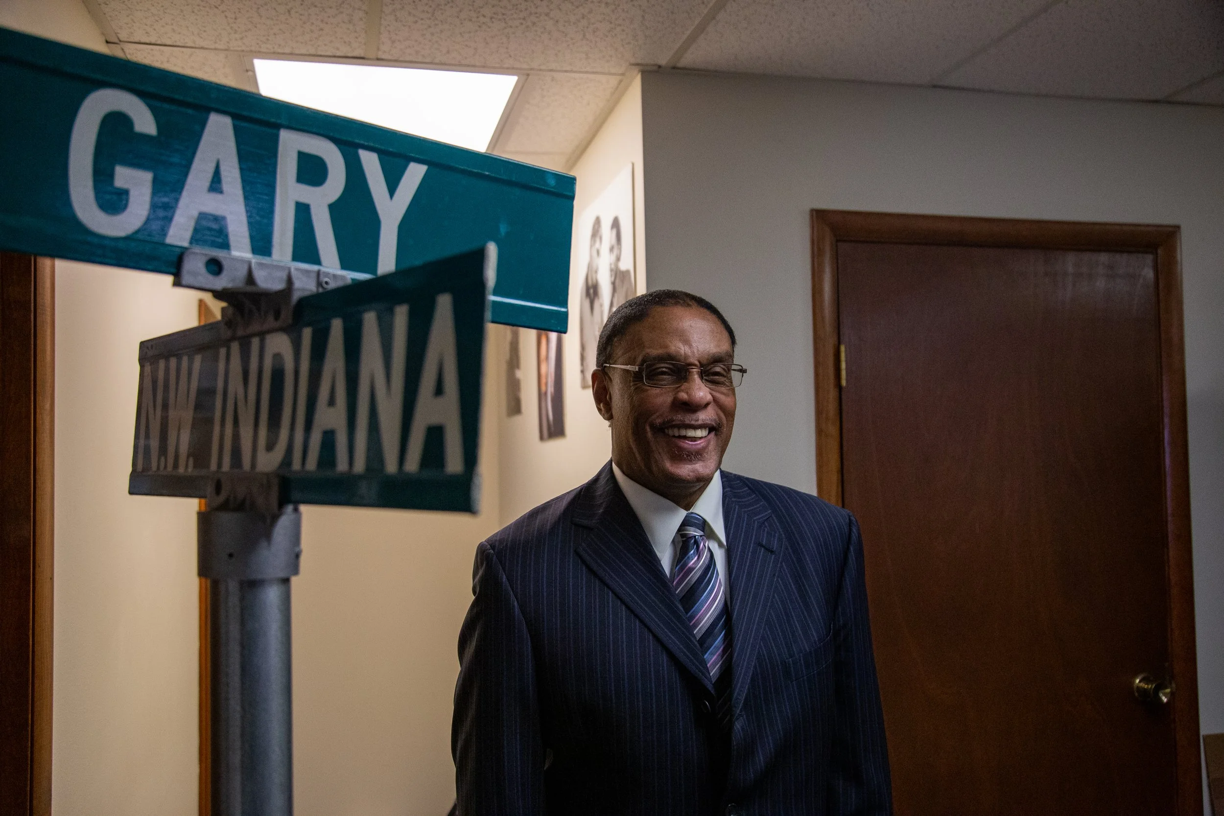 A man in a suit smiling, standing inside an office, with street signs for Gary and New Indiana in the foreground.